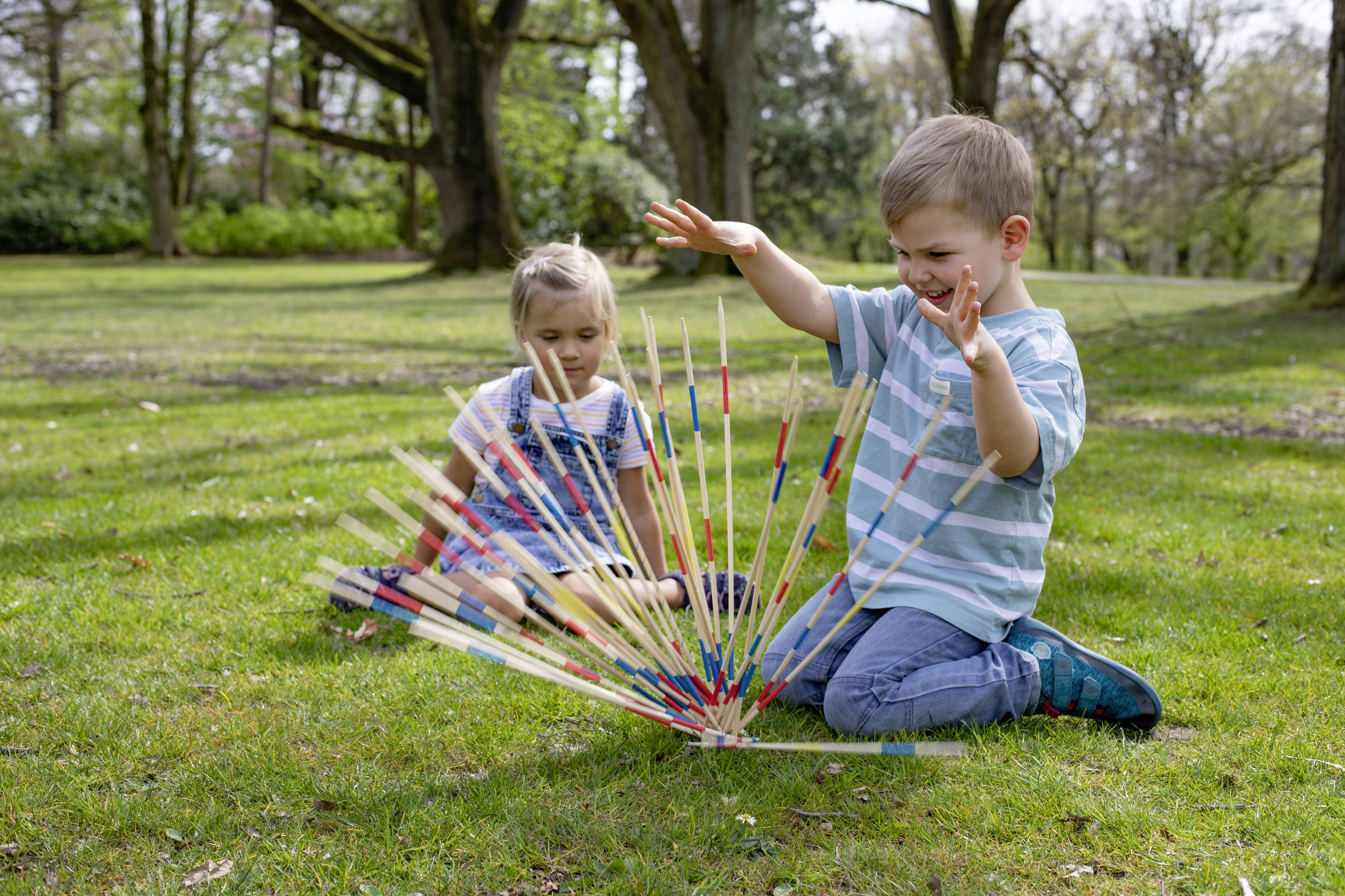 Dwoje dzieci gra w parku w Mikado. Jedno próbuje podnieść patyczki, podczas gdy drugie przygląda się. Otaczają ich drzewa.