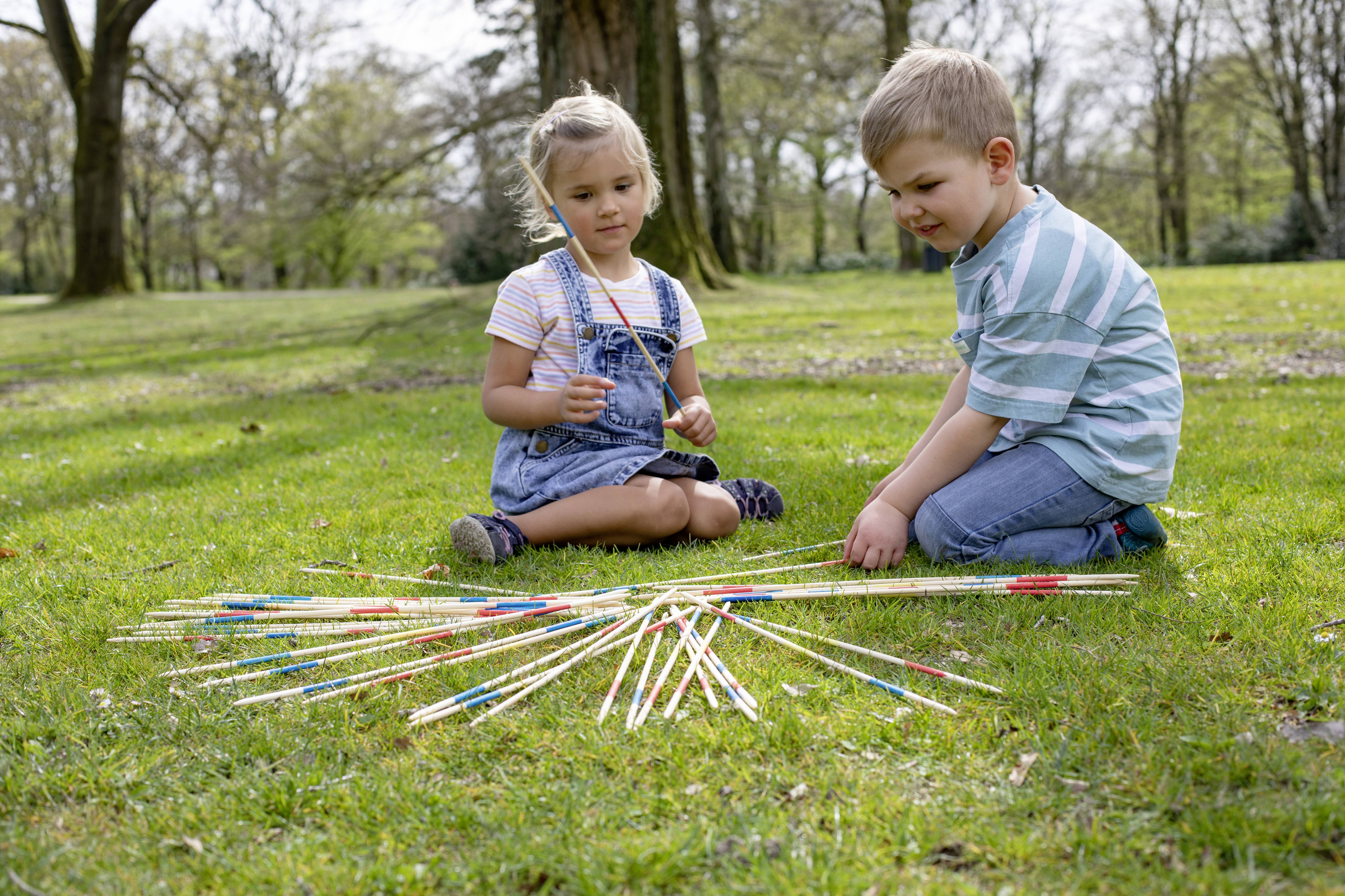 Dwoje dzieci gra na dworze w mikado na łące. Dziewczynka trzyma patyk, podczas gdy chłopiec siedzi i obserwuje. W tle widać drzewa.