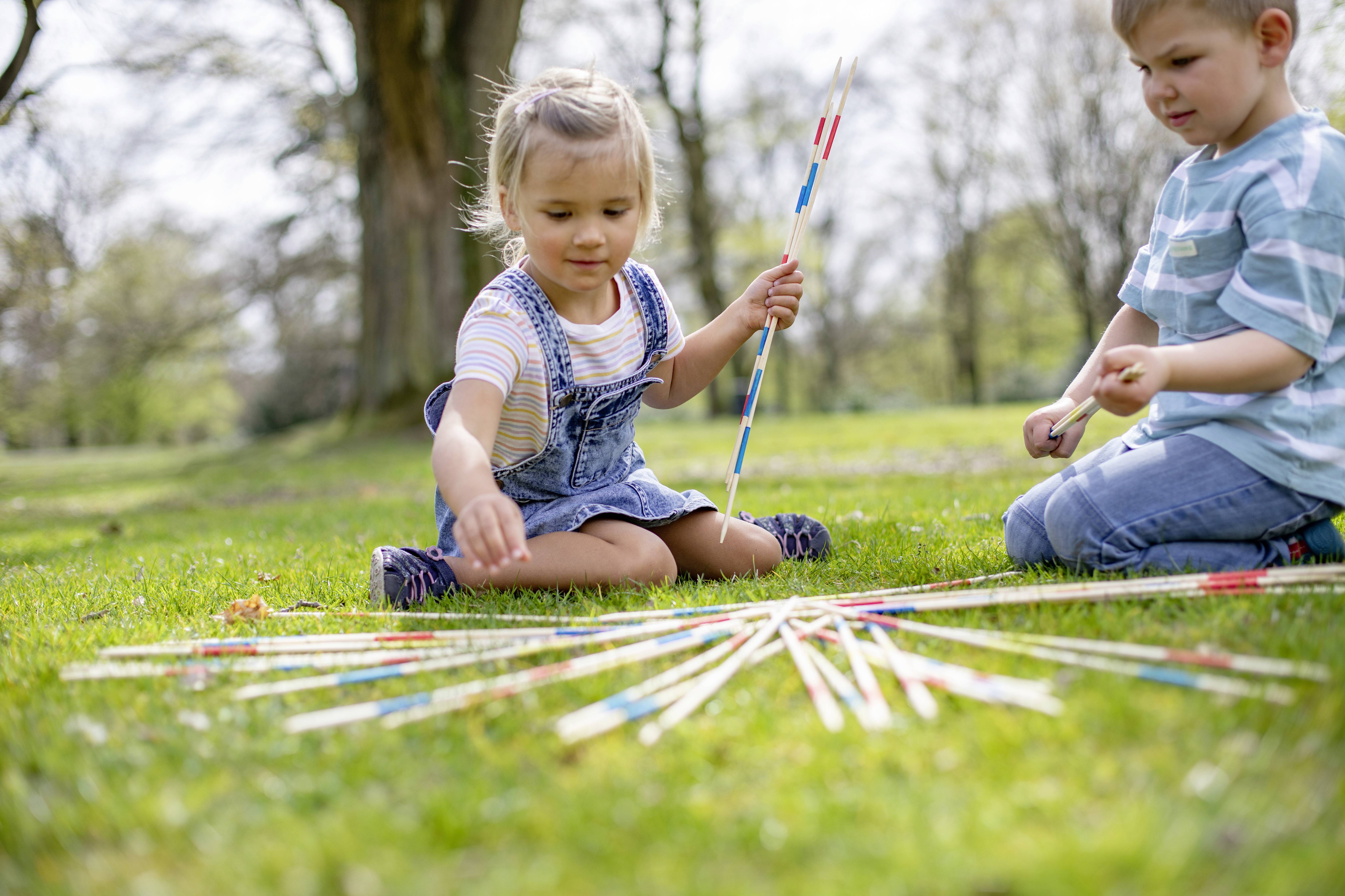 Dwoje dzieci bawi się na łące w Mikado, otoczonych drzewami. Dziewczynka trzyma patyk, podczas gdy chłopiec wybiera jeden.