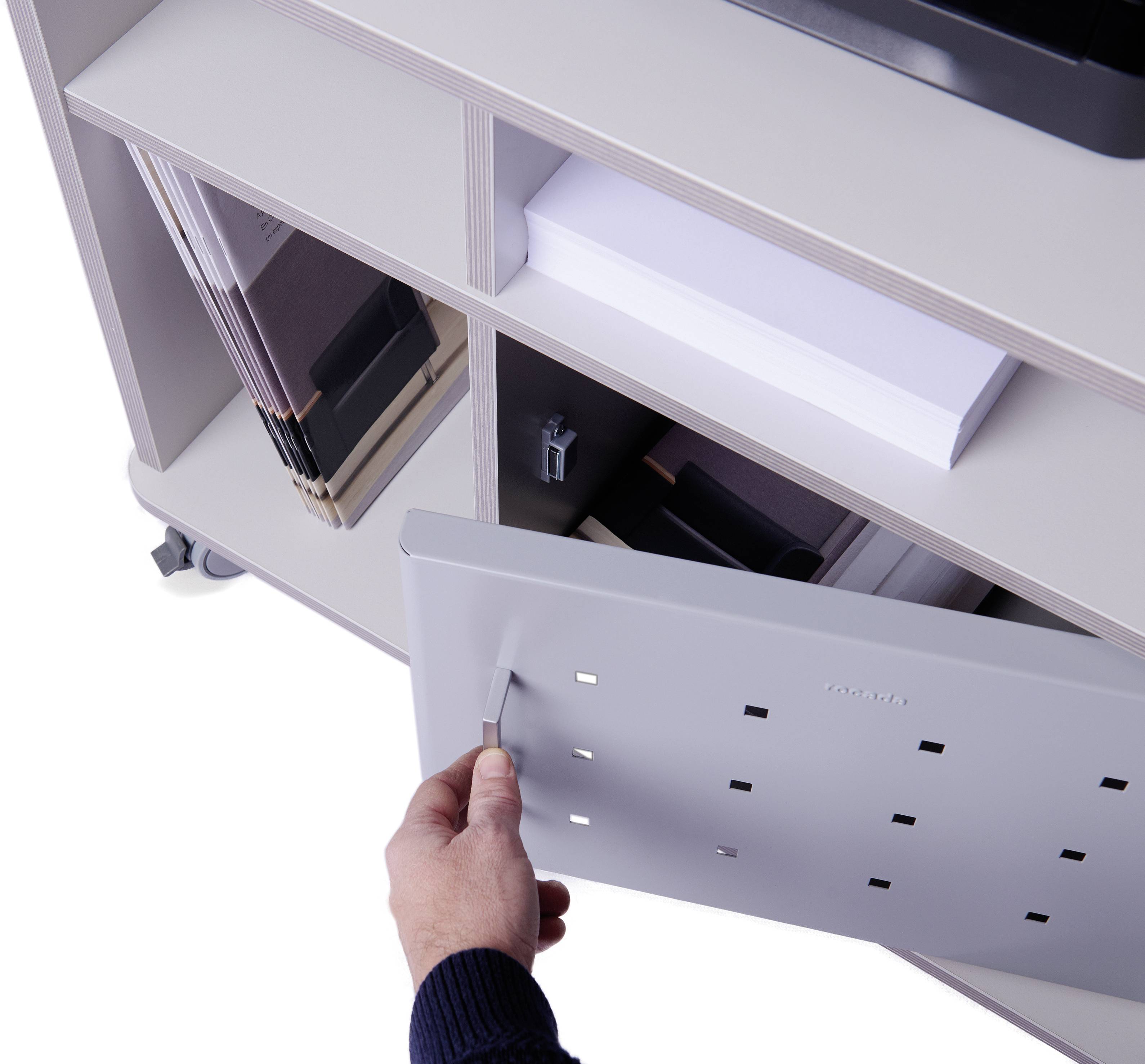 Ręka otwiera białą szufladę kontenera biurowego. W przegródce leżą segregatory i spinacze do papieru.