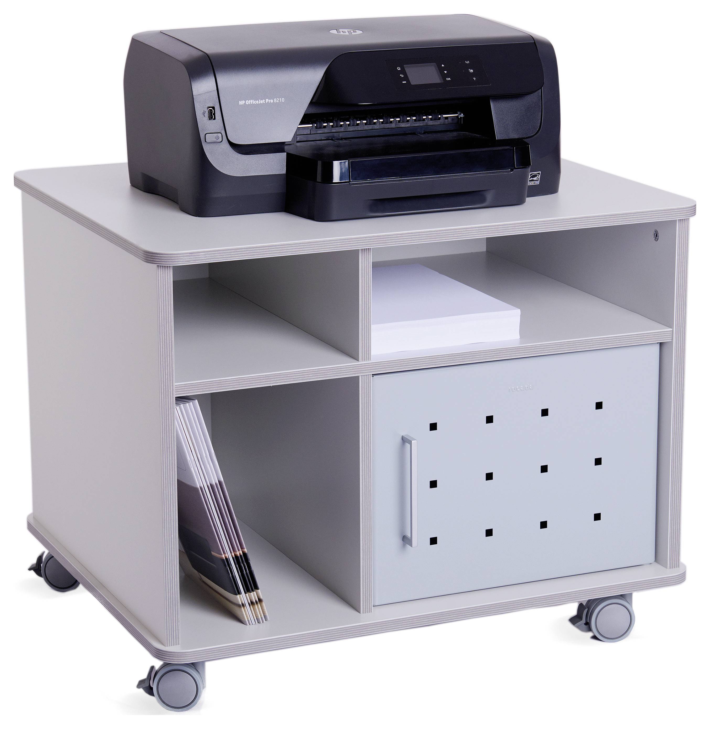 Drukarka stoi na szarym, ruchomym szafce z przegródkami. Na górze leżą stosy papieru, na dole znajdują się prospekty i drzwiczki.