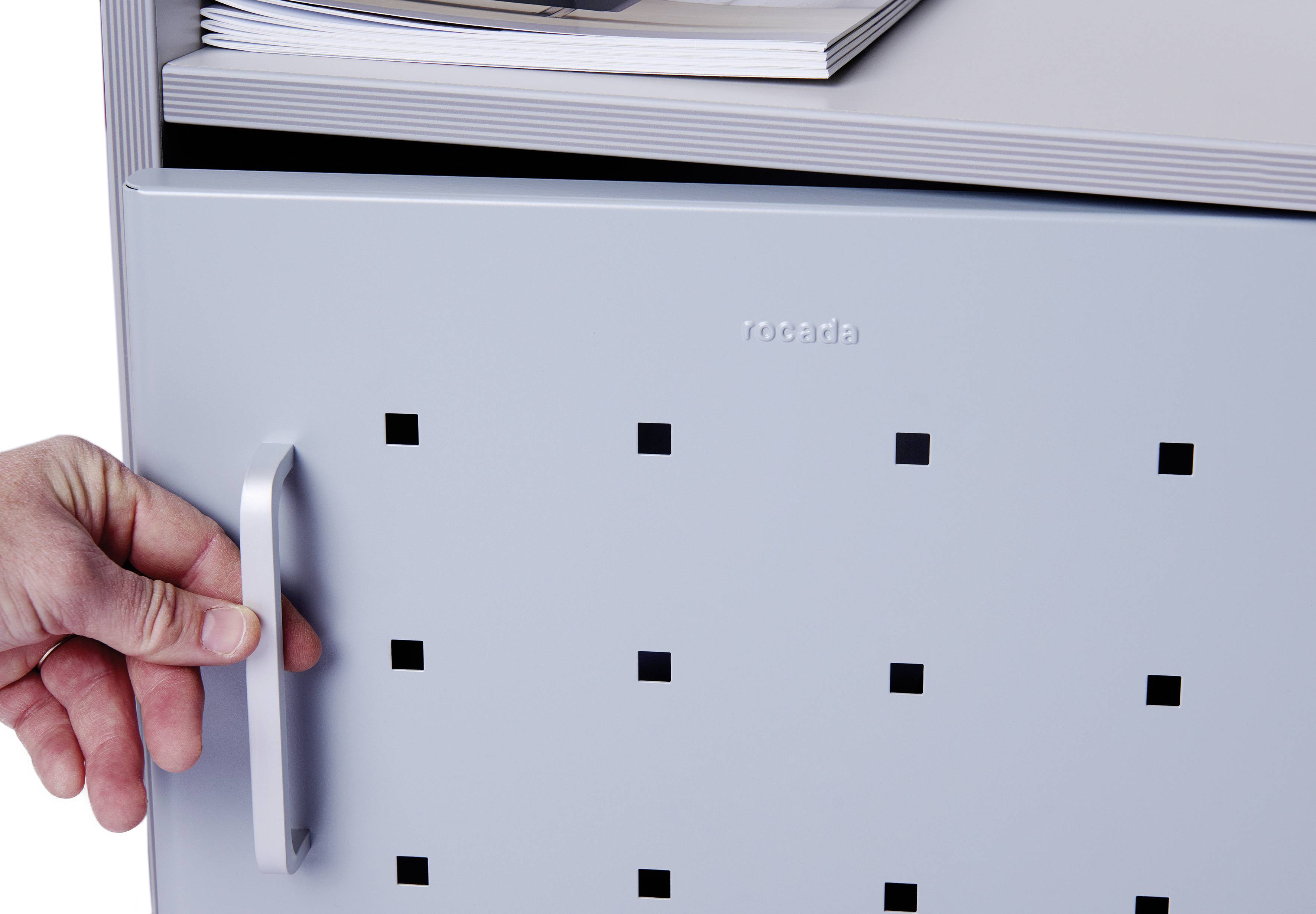 Ręka pociąga za kwadratowo dziurkowany uchwyt drzwi szarej metalowej szafki. Na górze leżą czasopisma.
