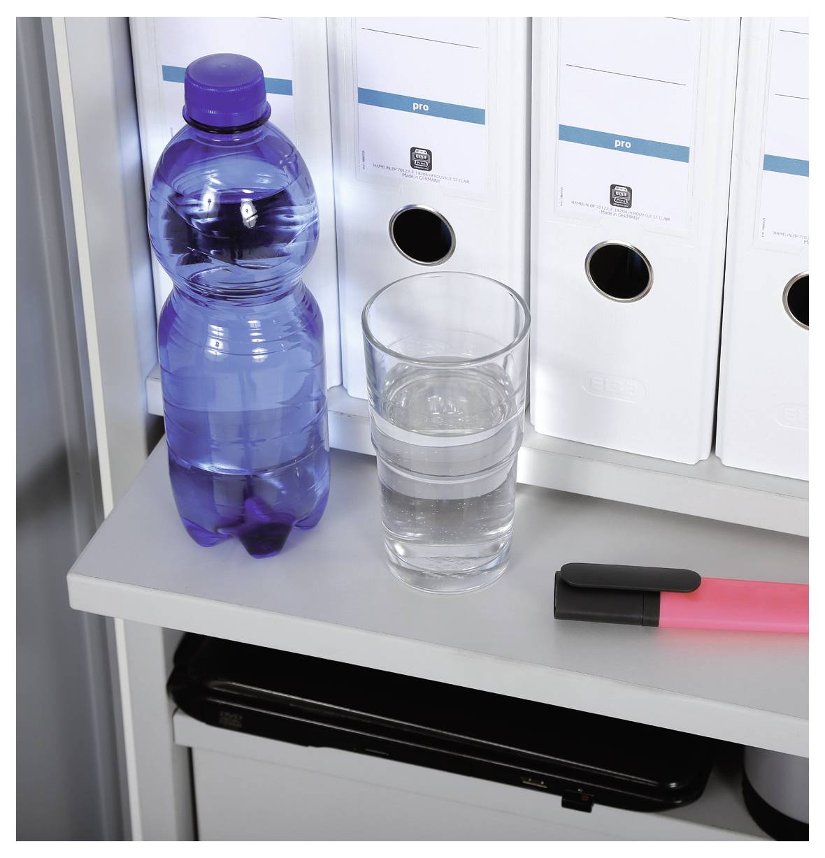 Na półce stoją: do połowy napełniona butelka wody, szklanka z czystą wodą, różowy zakreślacz i białe segregatory.