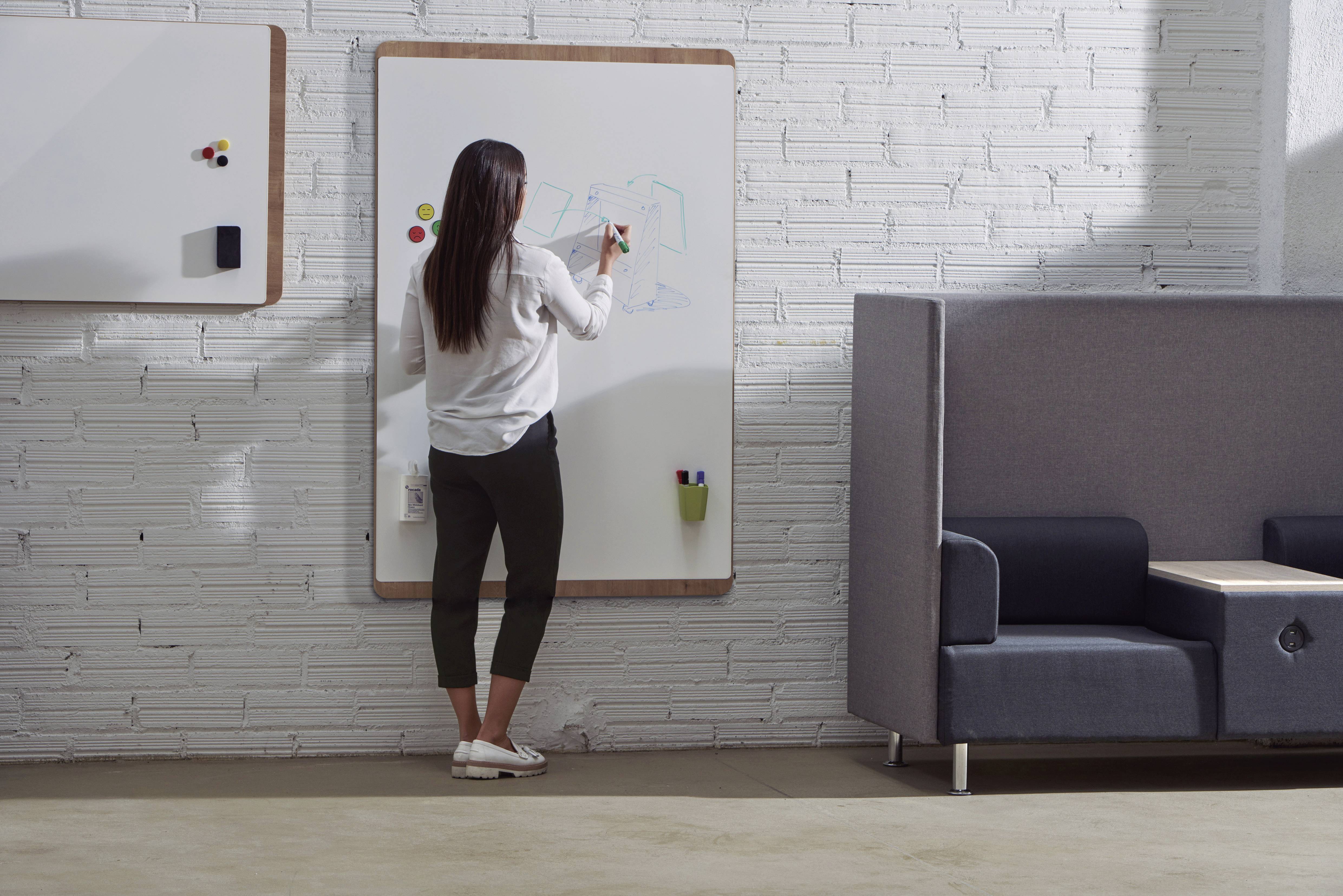 Kobieta rysuje na tablicy w nowoczesnym biurze. W tle znajduje się kanapa i biała ściana z cegły.