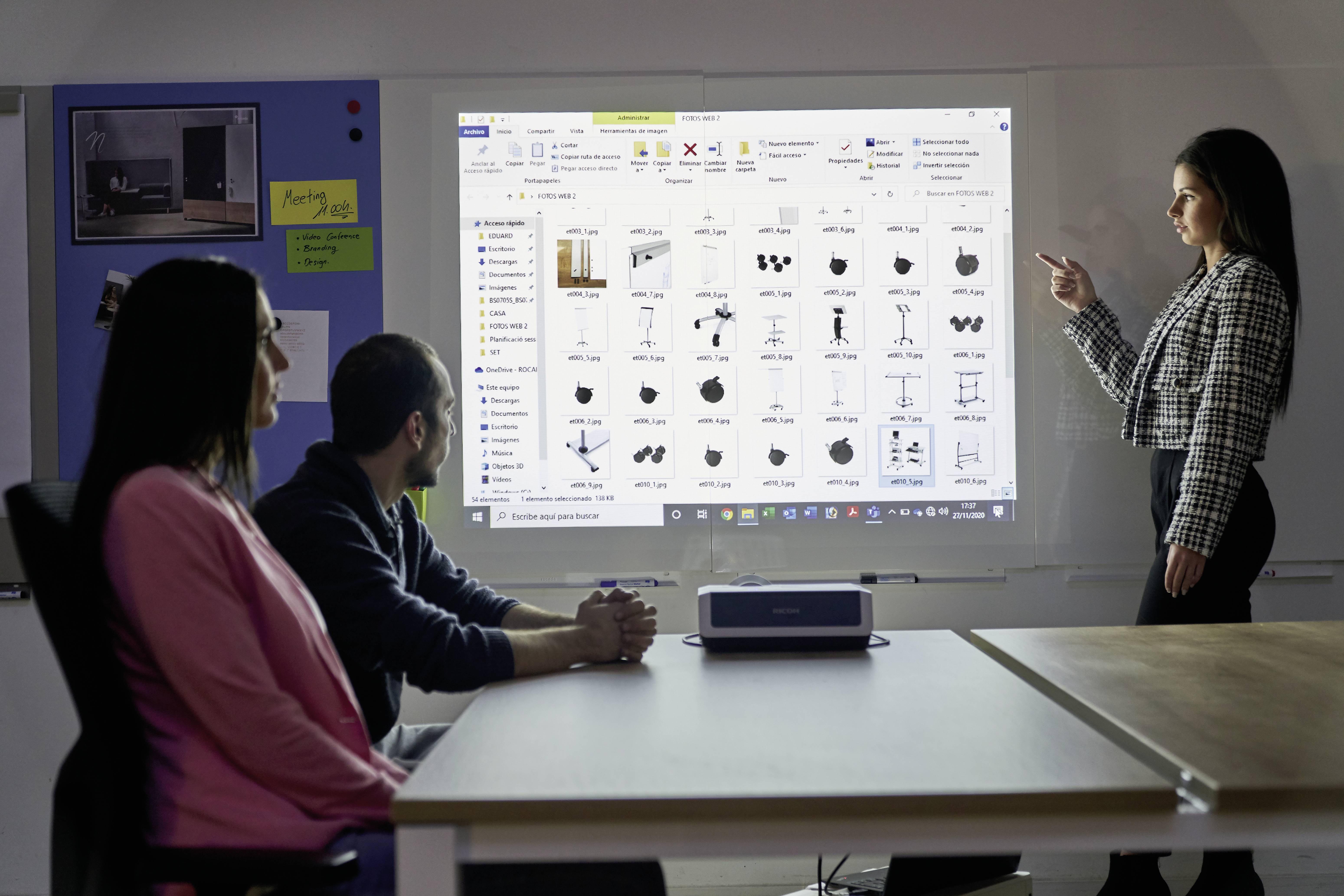 Kobieta stoi i prezentuje, podczas gdy dwie osoby siedzą przy stole i słuchają, a w tle na ekranie wyświetlane są obrazy.