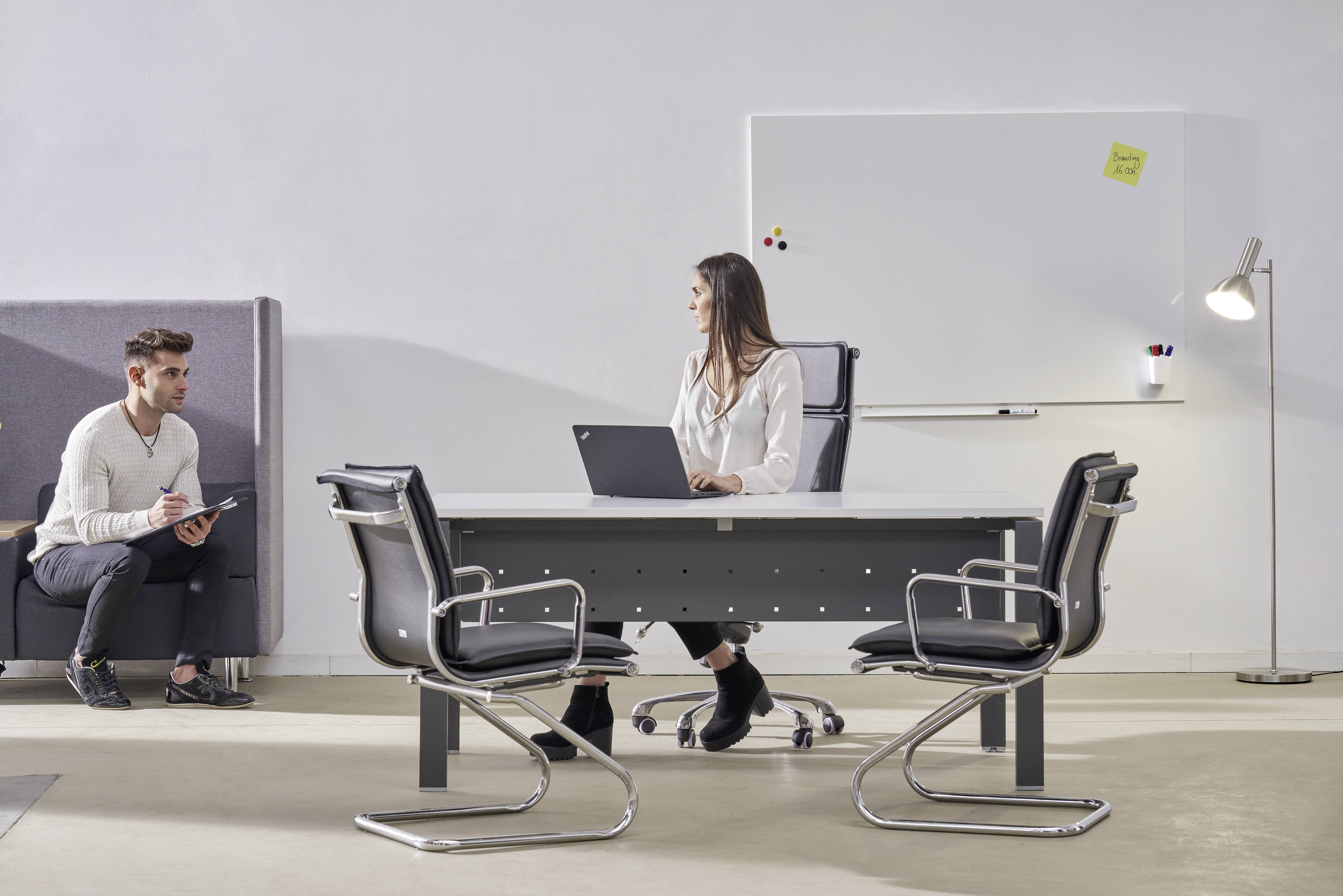 Dwie osoby w nowoczesnym biurze. Jedna siedzi przy biurku z laptopem, druga na kanapie, trzymając tablet. W tle tablica whiteboard.