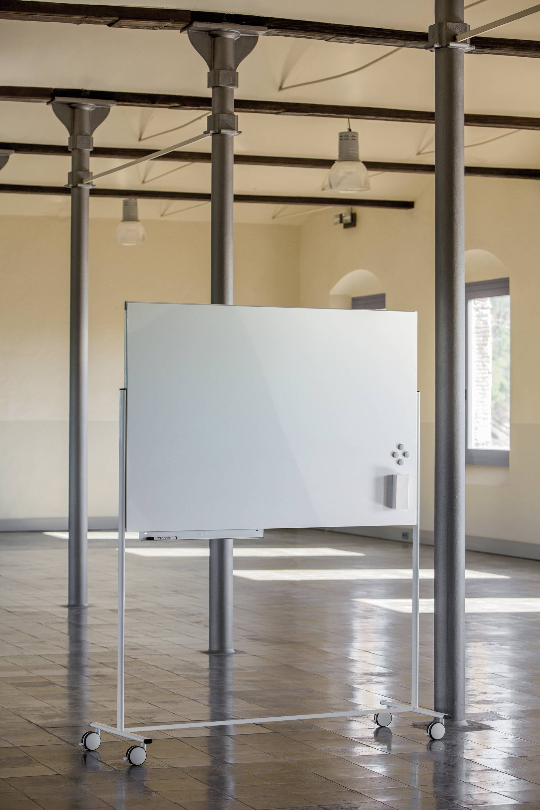 Mobilna tablica na kółkach stoi w dużej, jasnej przestrzeni z podłogą wyłożoną płytkami i kolumnami. Idealna do prezentacji lub warsztatów.