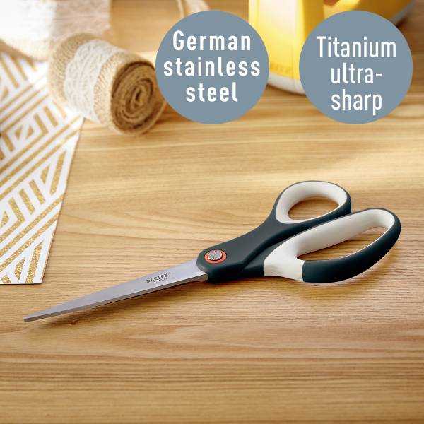 Nożyczki leżą na stole pośród rolek materiału. Napisy wskazują na niemiecką stal nierdzewną i ultrawytrzymałe tytanowe ostrze.