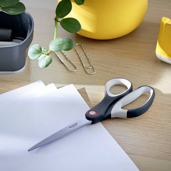 Nożyczki leżą na biurku obok dwóch spinaczy, liści roślin, żółtego pojemnika i kartki papieru.