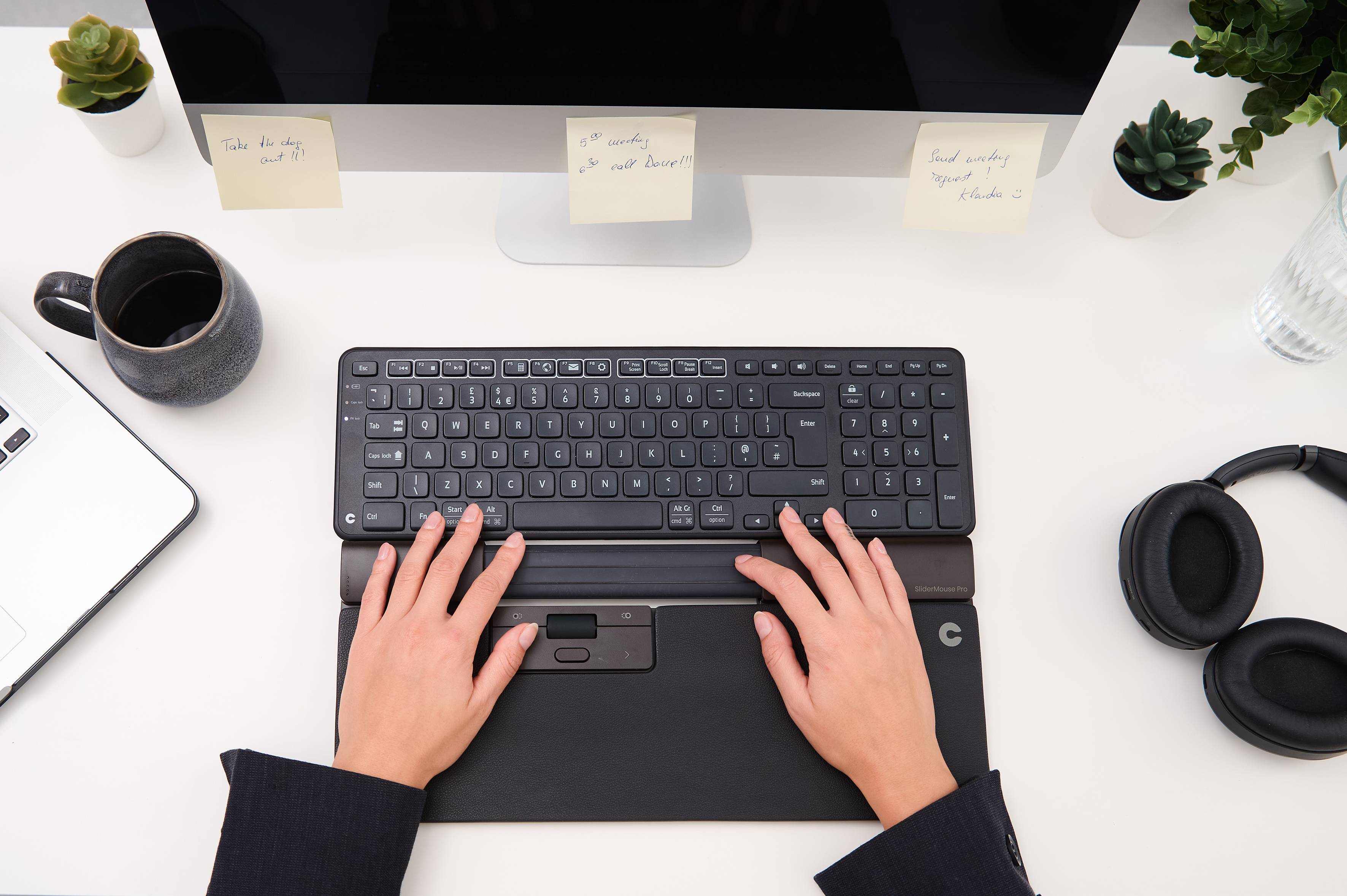 Osoba pisze na czarnej klawiaturze, na biurku z laptopem, kubkiem, słuchawkami, monitorem i karteczkami samoprzylepnymi.