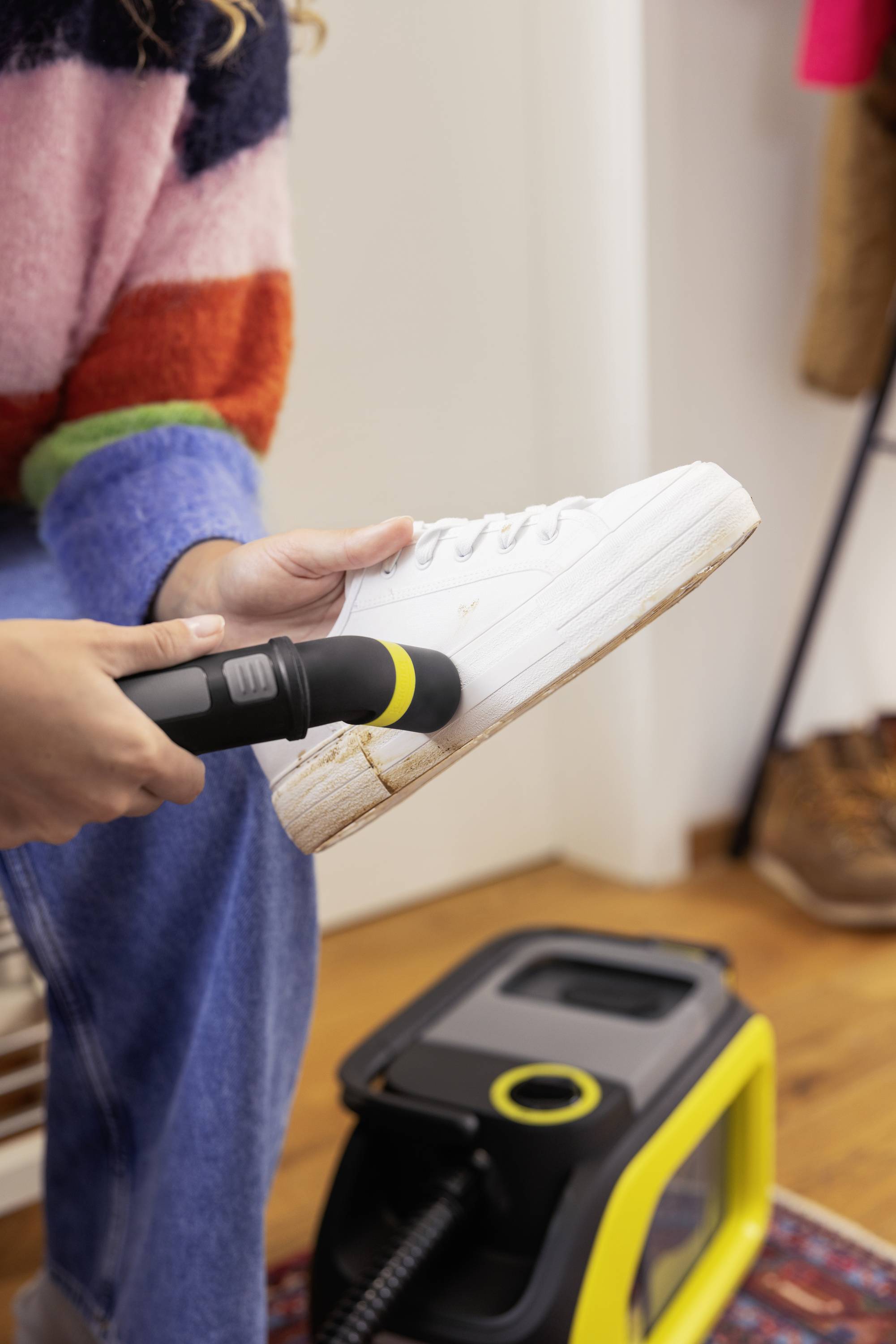 Osoba czyści białego buta sportowego małą szczoteczką i parownicą. But sportowy ma widoczne plamy i zabrudzenia.