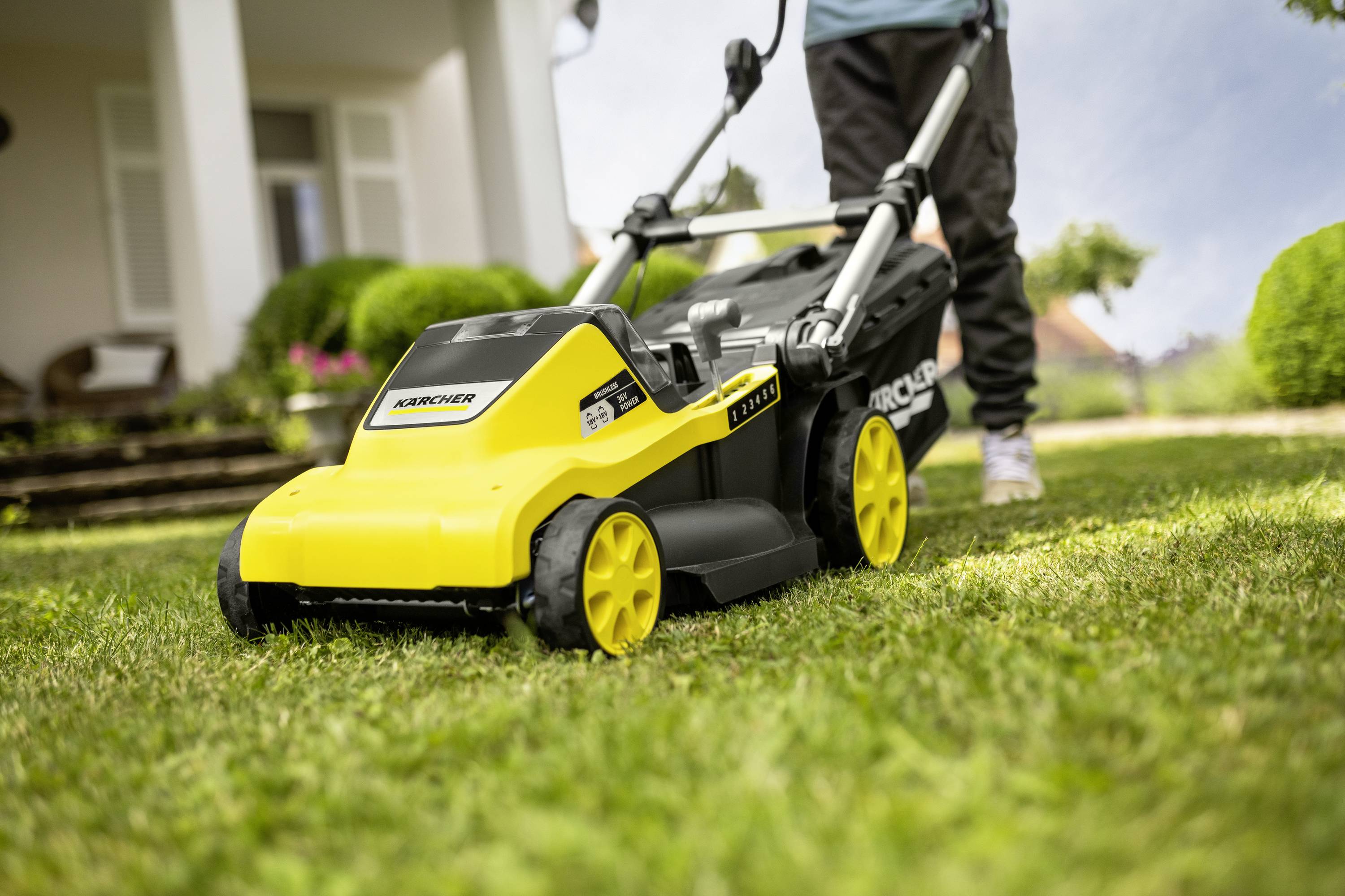Osoba przesuwa żółtą kosiarką po świeżo skoszonym trawniku przed domem z białymi kolumnami i zielonymi krzewami.