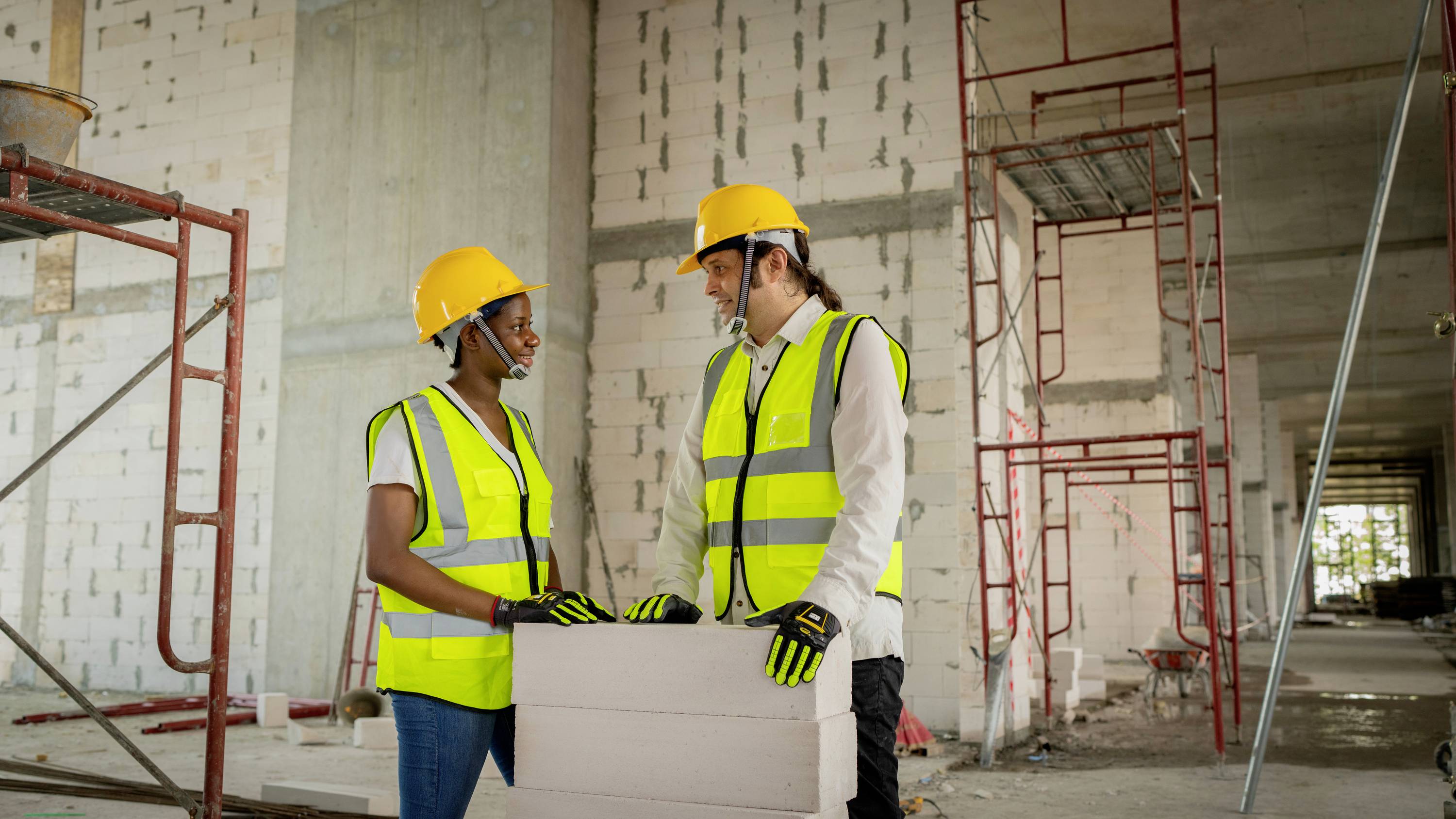 Dwaj robotnicy budowlani w kaskach ochronnych i kamizelkach ostrzegawczych rozmawiają w niedokończonym budynku. Trzymają w rękach plany. W tle widoczne są rusztowania.