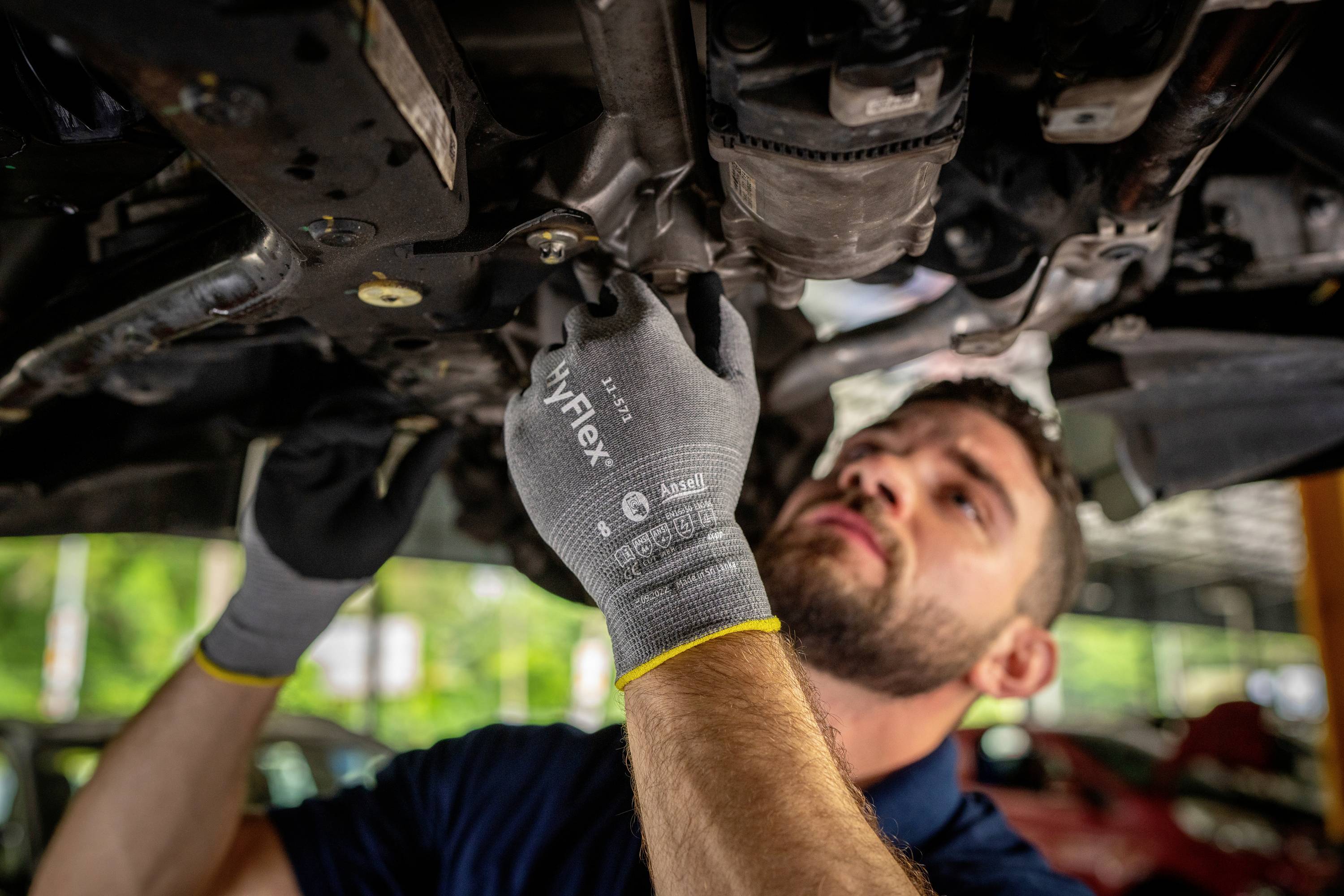 Mechanik naprawia pojazd od spodu, ma na sobie szare rękawice ochronne, twarz wyrażająca skupienie, w tle widoczne są samochody.