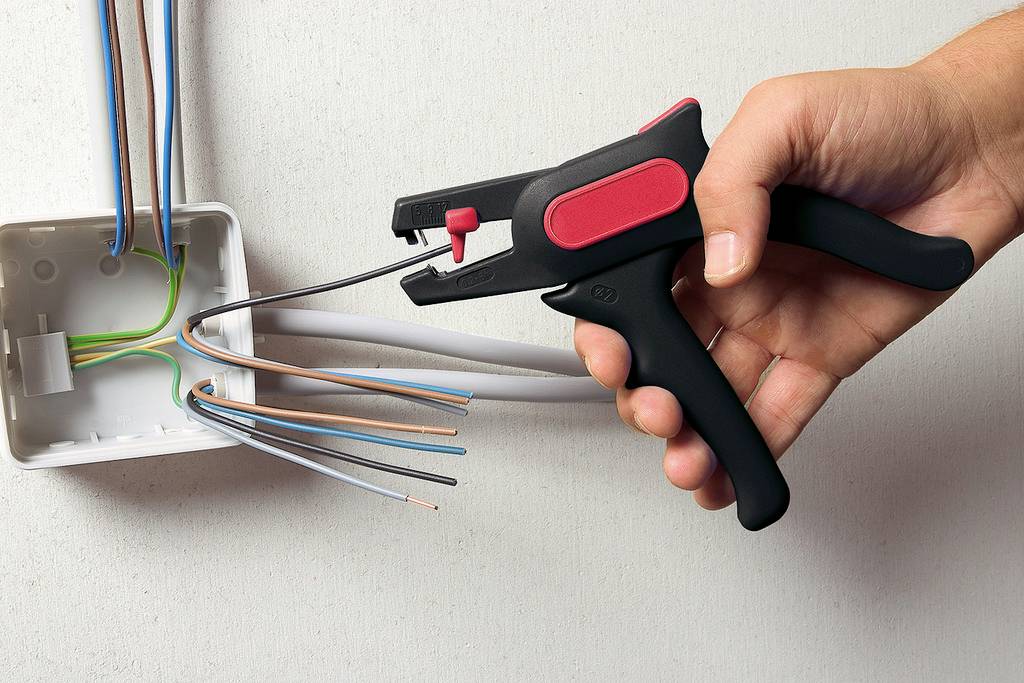 Jedna ręka trzyma narzędzie do zdejmowania izolacji i usuwa izolację z kilku kabli wystających z puszki ściennej.