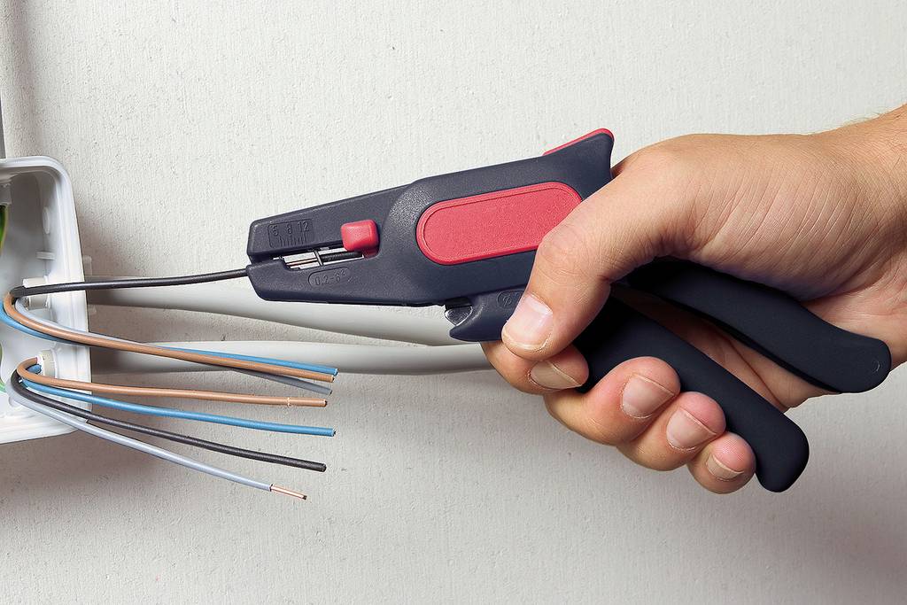 Ręka trzyma obcinacz izolacji i usuwa izolację z kilku przewodów elektrycznych wystających z puszki ściennej.