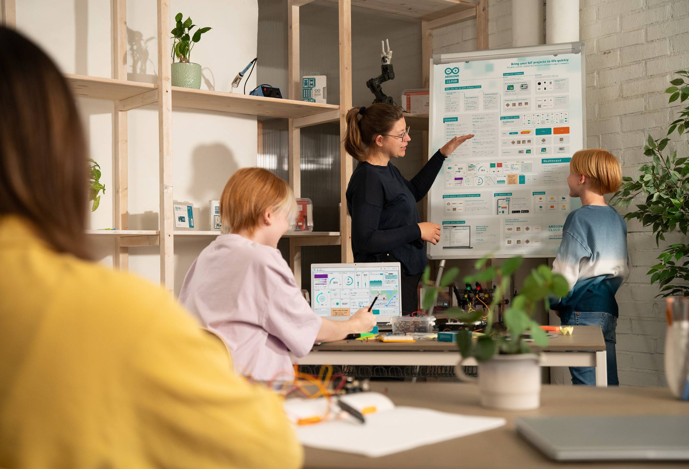 Nauczycielka pokazuje dwóm uczniom plakat w klasie. Uczniowie uważnie przyglądają się plakatowi, siedząc przy stole.