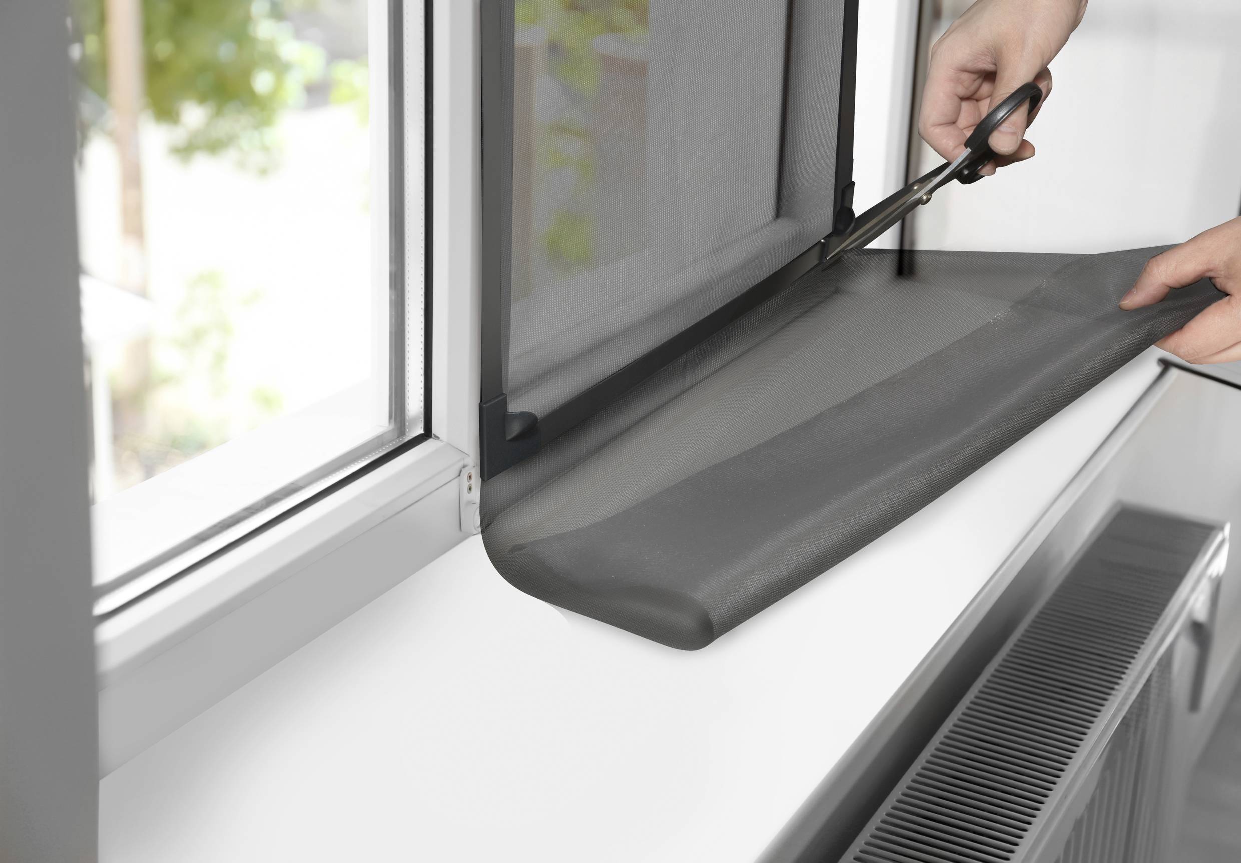 Osoba przycinająca nożyczkami siatkę przeciw owadom, aby dopasować ją do otwartego okna. W tle niewyraźnie widoczne są drzewa.
