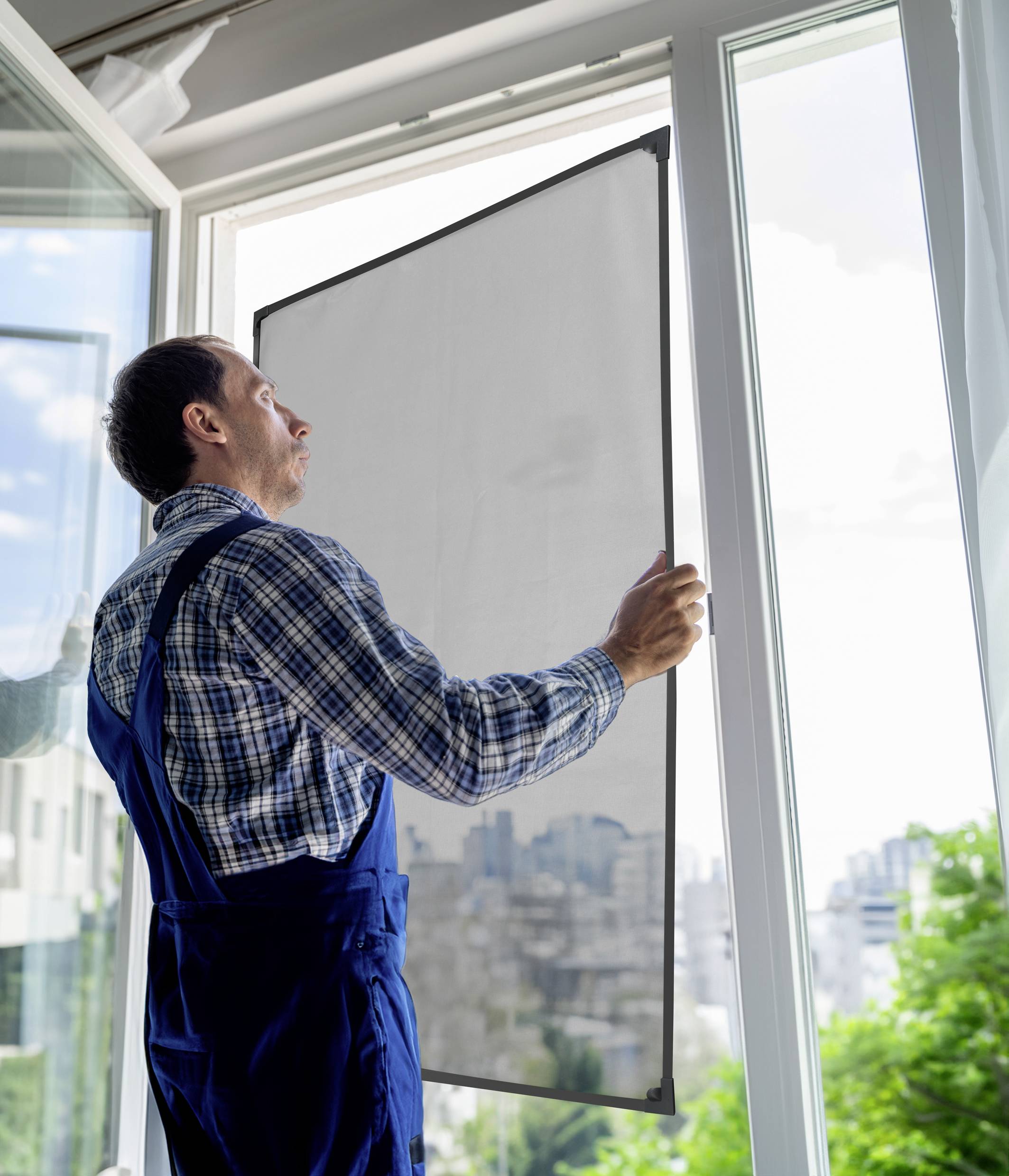 Mężczyzna w stroju roboczym montuje ramkę przeciw owadom w otwartym oknie. W tle widać budynki i drzewa.