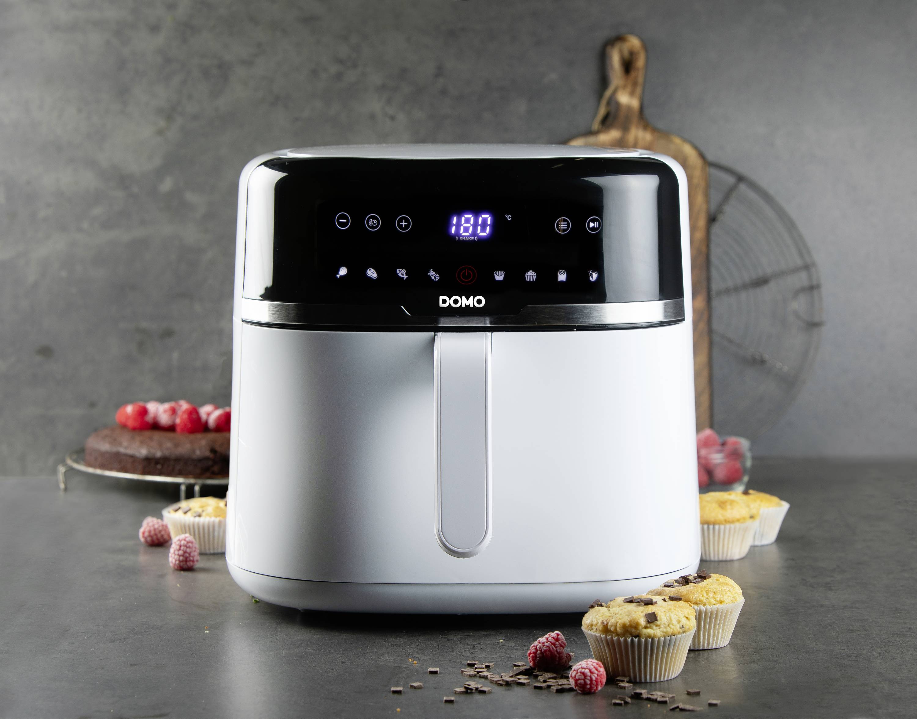 Biała frytownica na gorące powietrze stoi na stole kuchennym. W tle widać drewnianą deskę oraz wypieki, takie jak muffiny i ciasto.