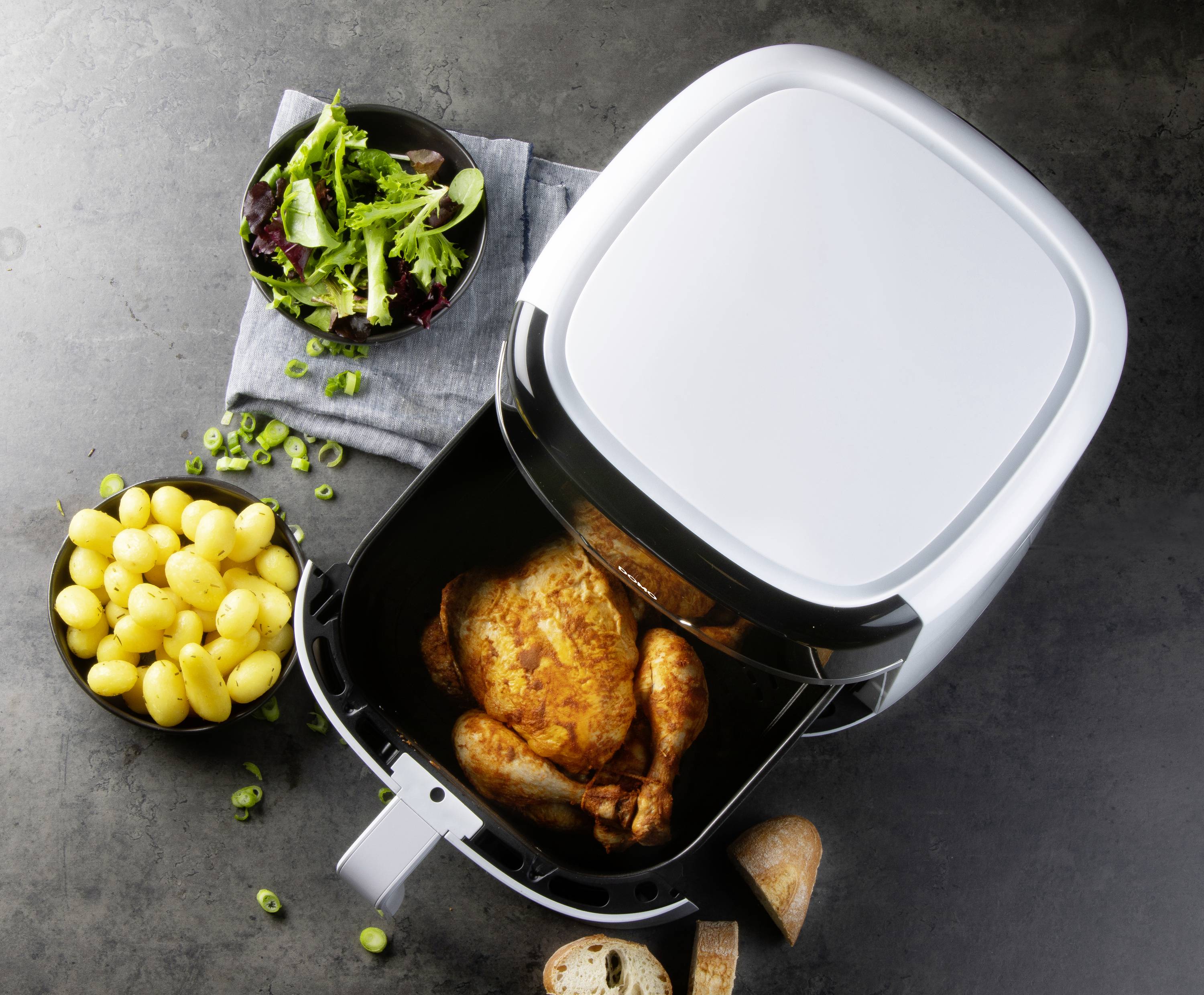 Frytkownica powietrzna z usmażonym kurczakiem, obok miseczki z ziemniakami, sałatką i pokrojonym chlebem na szarym podłożu.