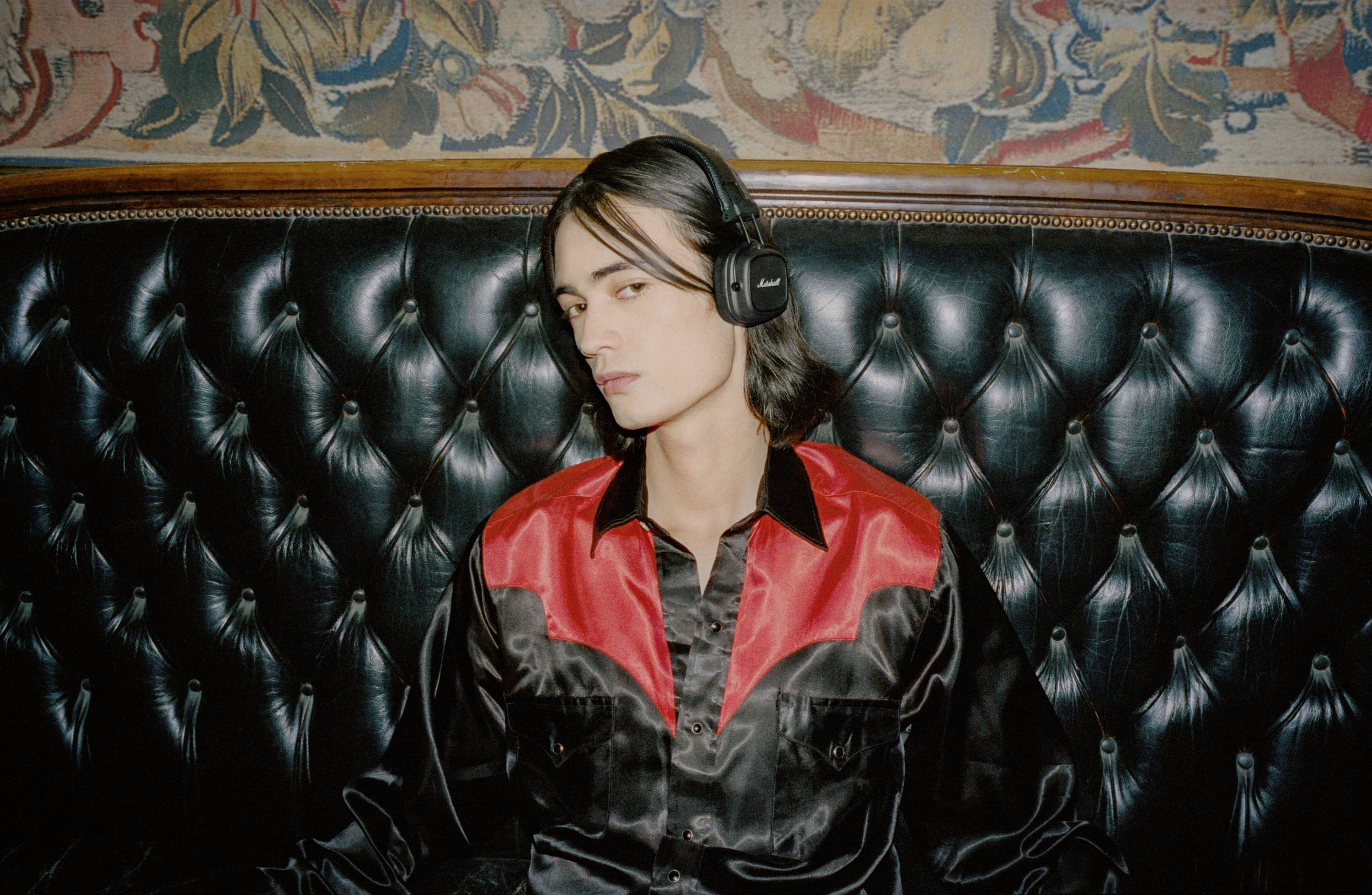 Osoba siedzi na skórzanej sofie z poduszkami, ubrana w czarno-czerwoną koszulę i czarne słuchawki. W tle widoczny jest kolorowy wzór.