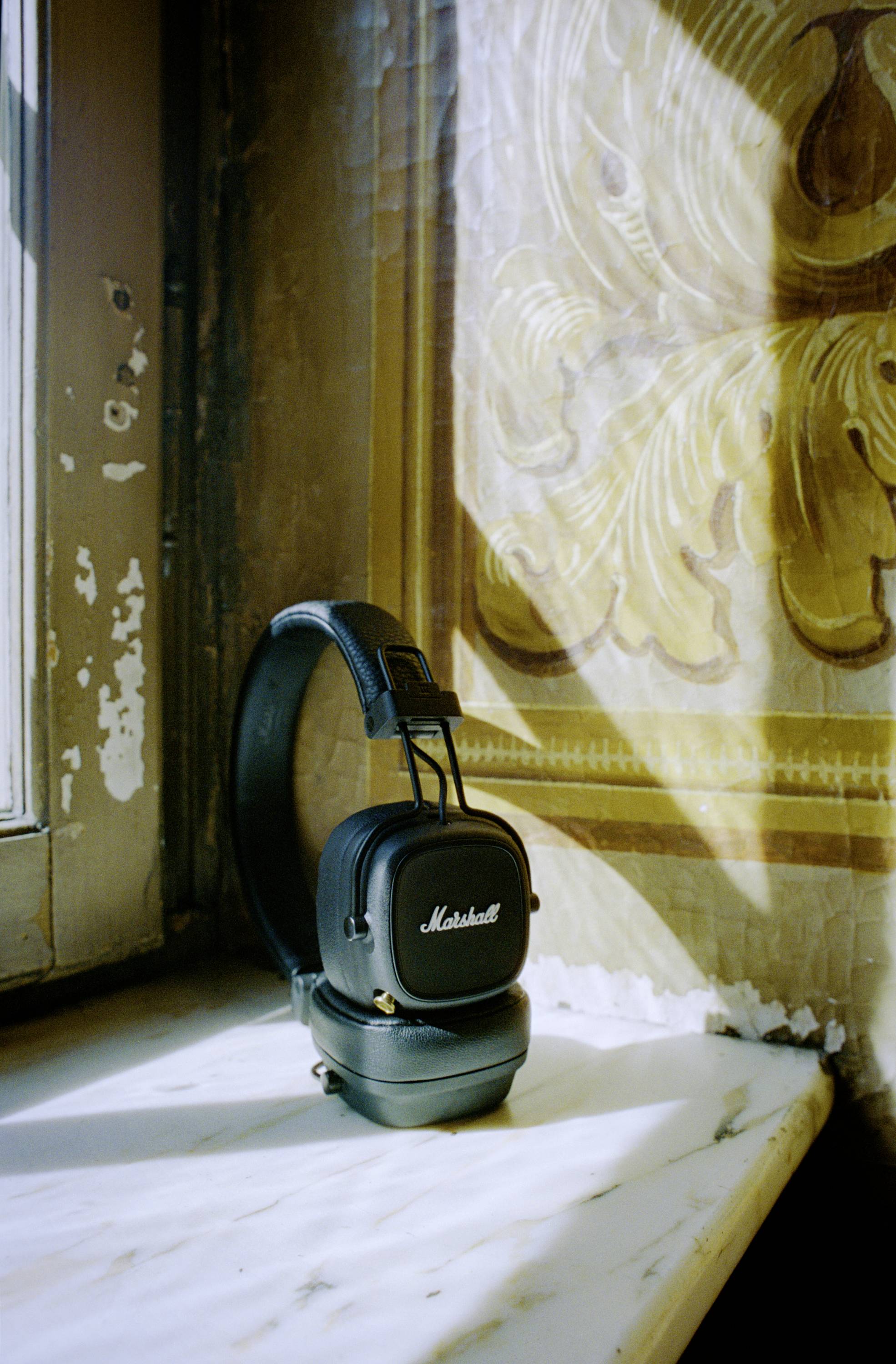 Czarny słuchawki z logo 'Marshall' leżą na parapecie w nasłonecznionym pokoju ozdobiony dekoracyjnymi malowidłami ściennymi.