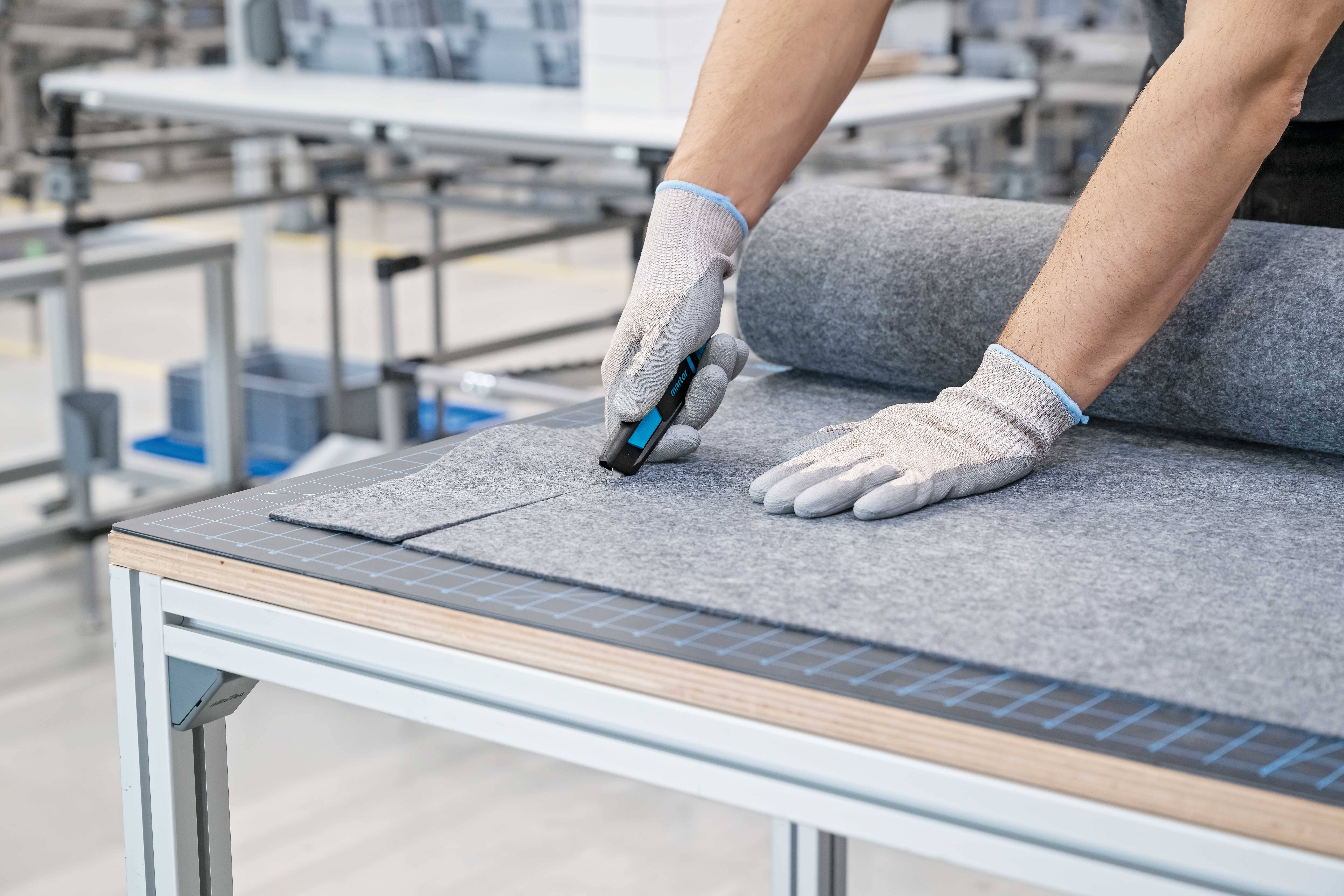 Osoba w rękawiczkach przycina duży, szary materiał wełniany narzędziem do cięcia na stole.