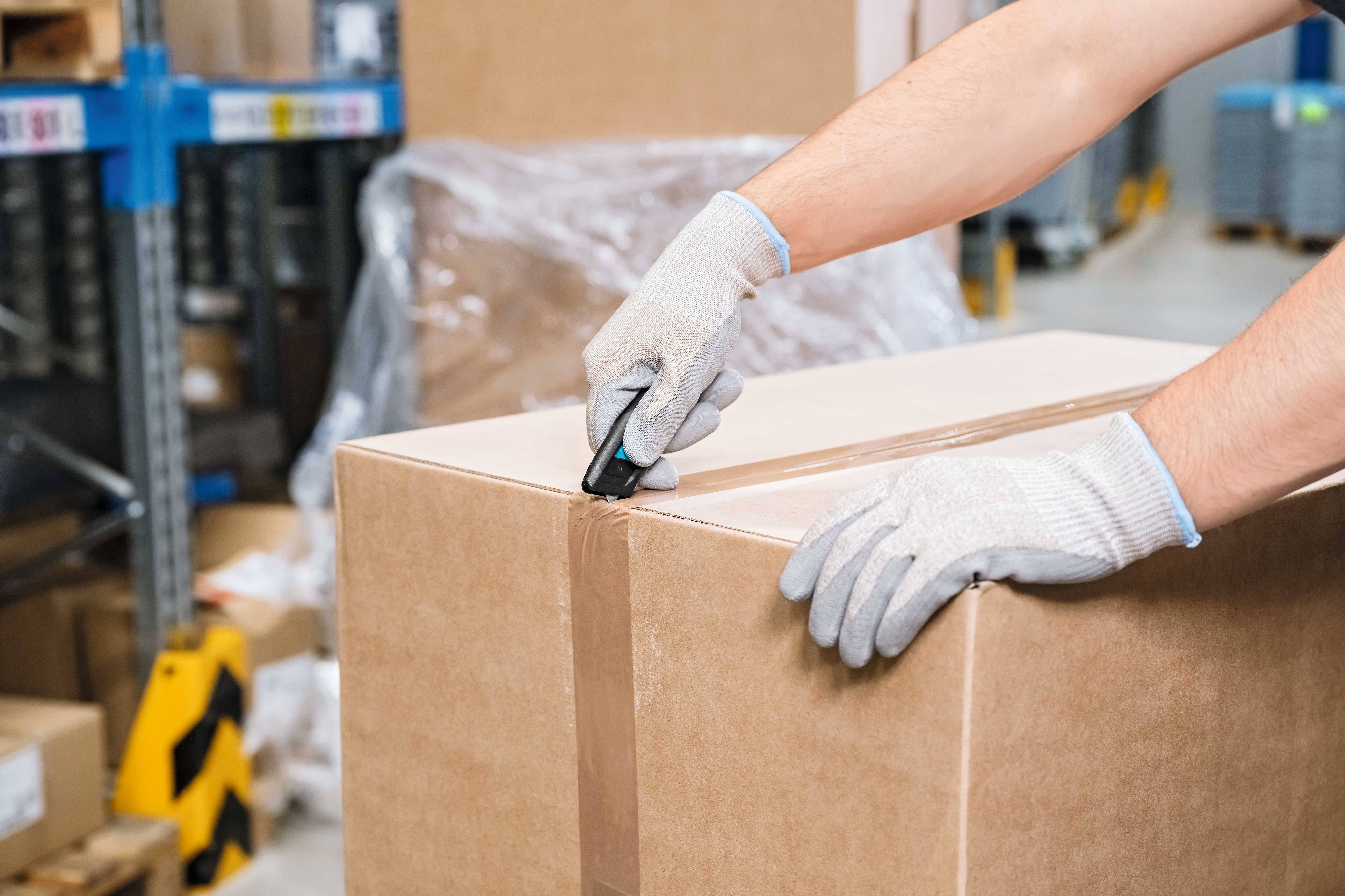 Osoba w rękawiczkach otwiera duży, zabezpieczony taśmą paczkę za pomocą noża monterskiego w magazynie.