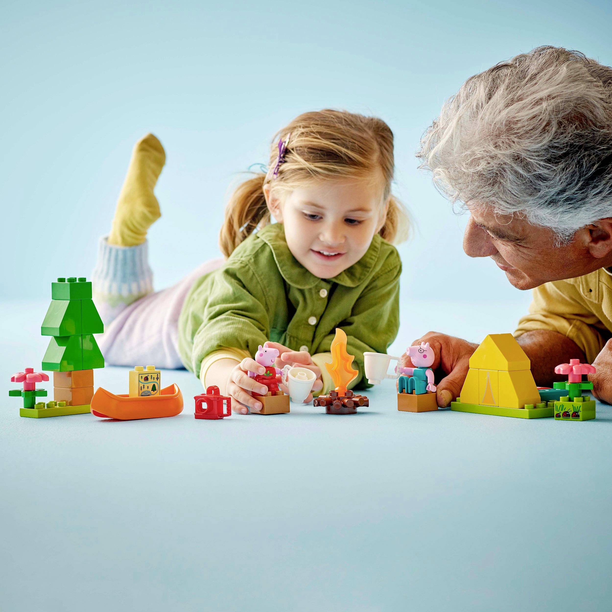 Starszy mężczyzna i dziecko bawią się kolorowymi klockami i figurkami na niebieskim podłożu. Sceny dotyczą przyrody i campingu.