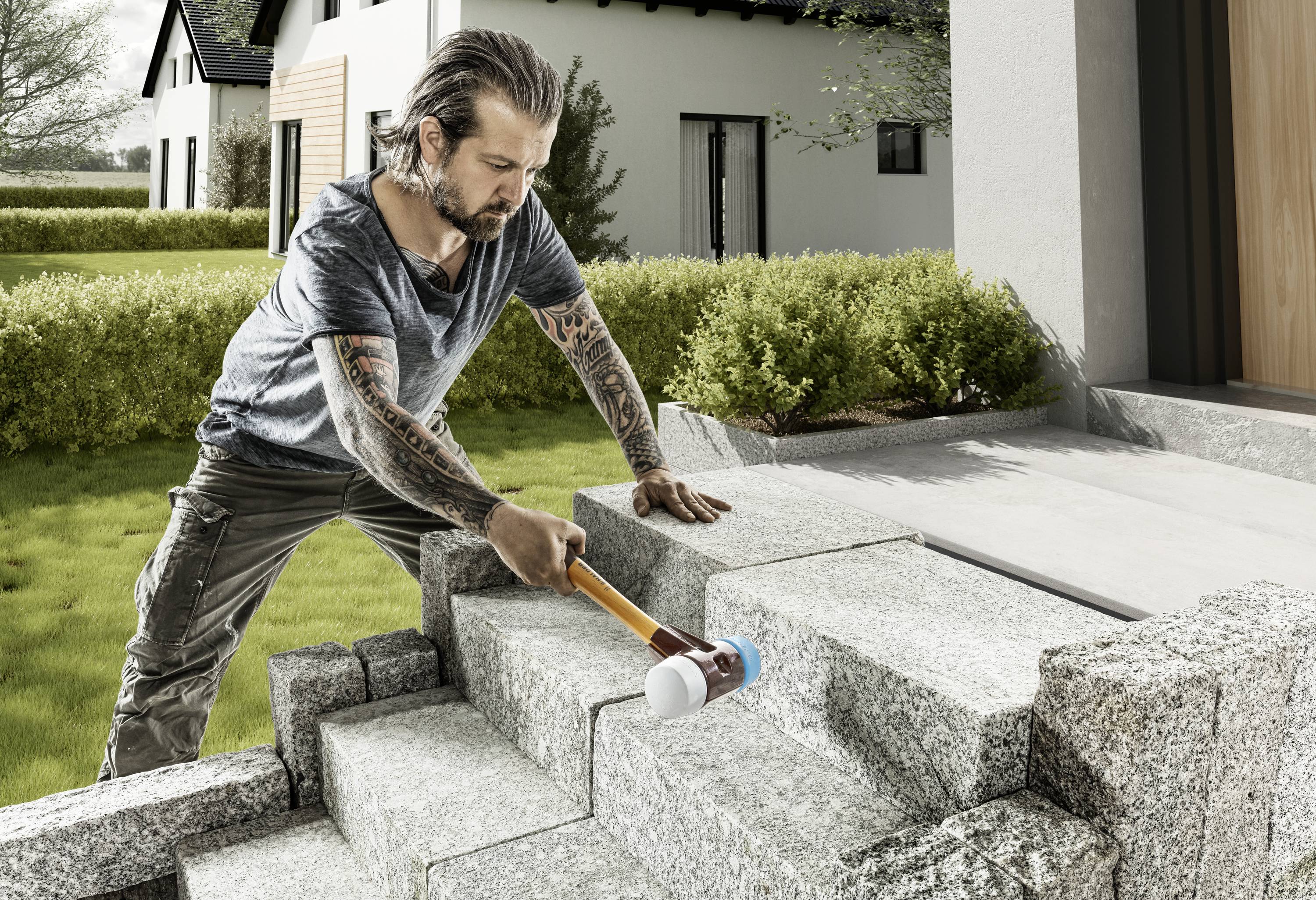 Mężczyzna z wytatuowanymi ramionami młotkuje kamienne schody nowoczesnego domu na wolnym powietrzu.