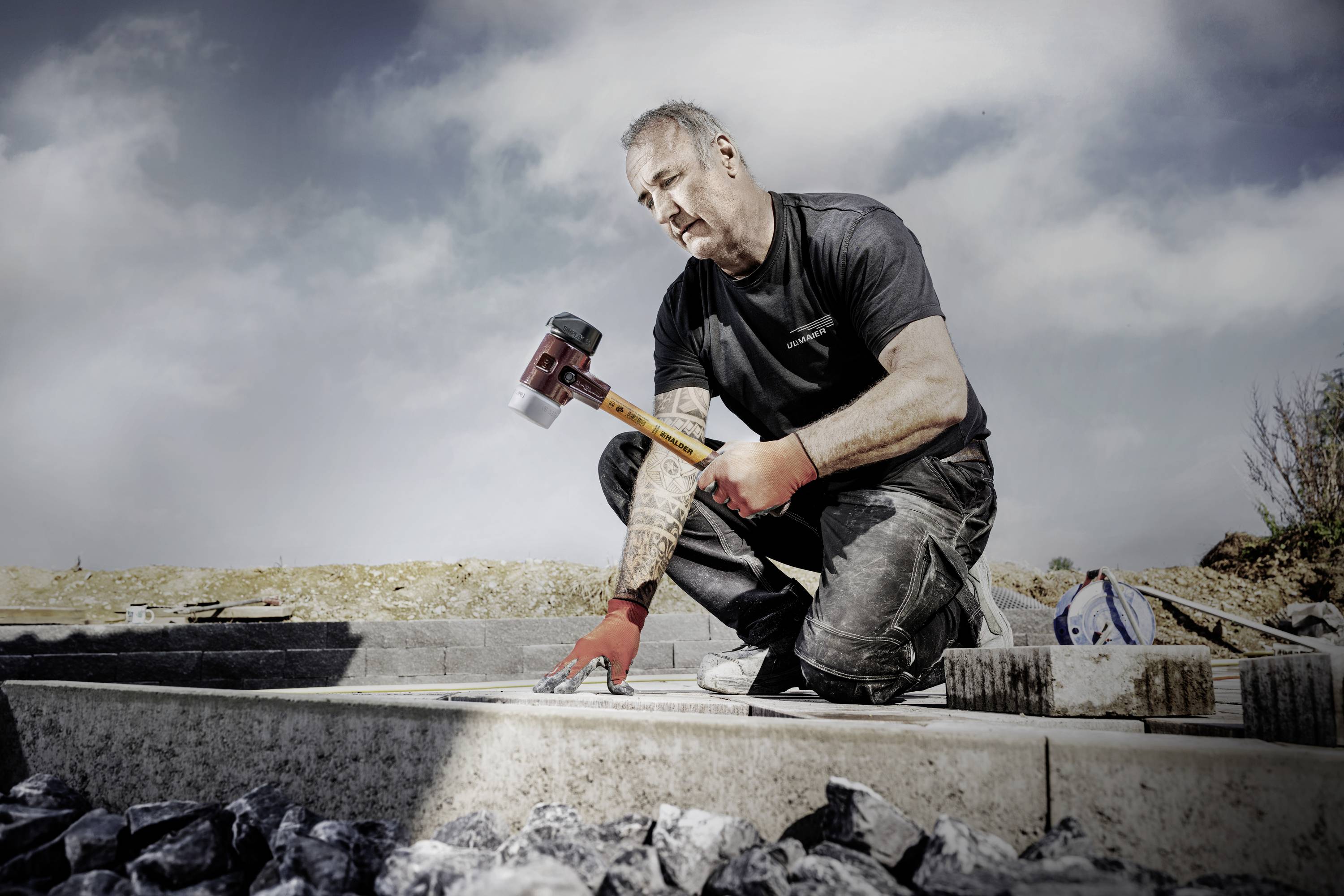 Robotnik w czerwonych rękawicach klęczy na budowie i obrabia kamień młotkiem, otoczony żwirowymi kamieniami.