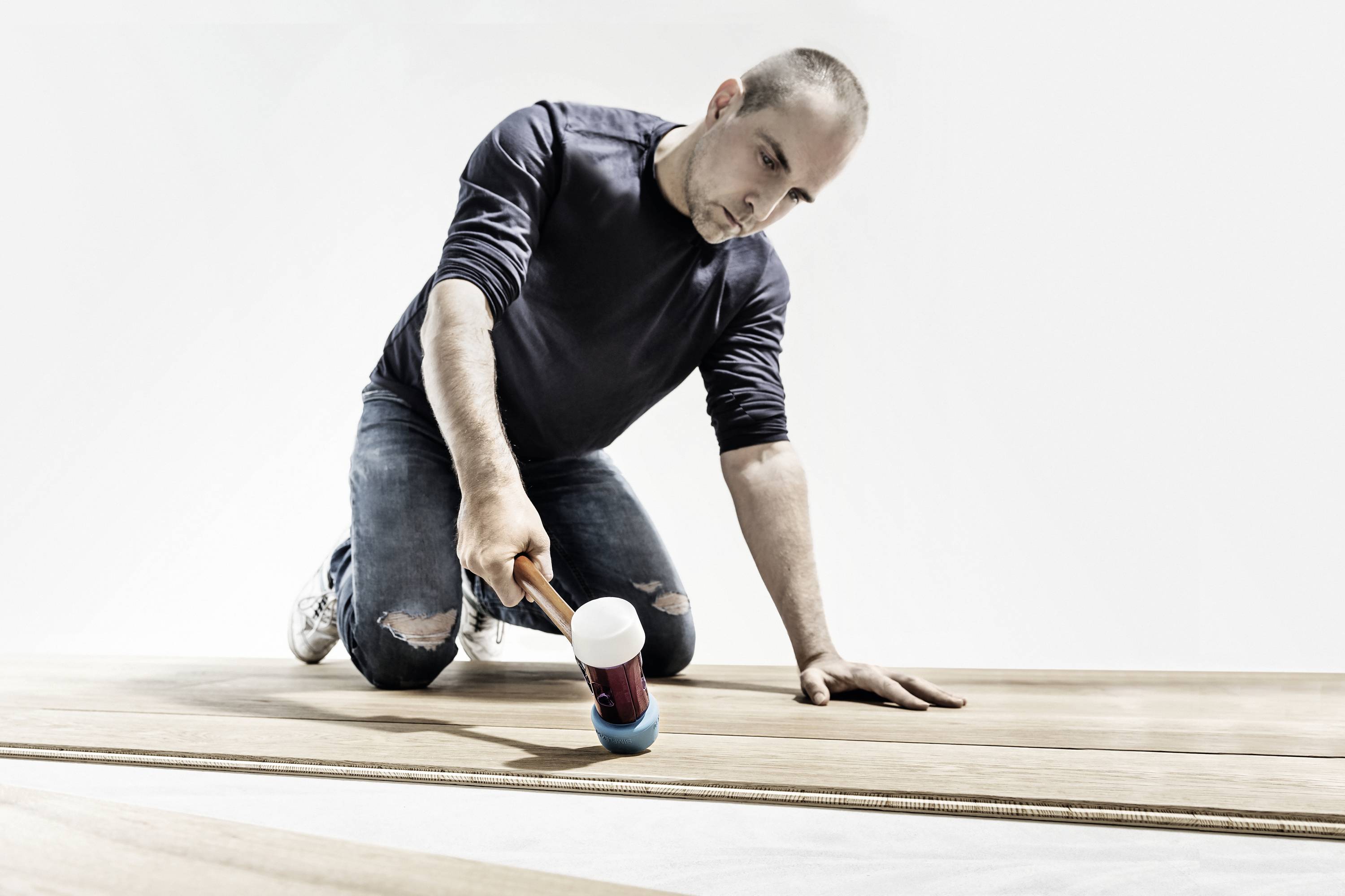 Mężczyzna klęczy na podłodze i instaluje drewnianą podłogę, używając młotka gumowego.