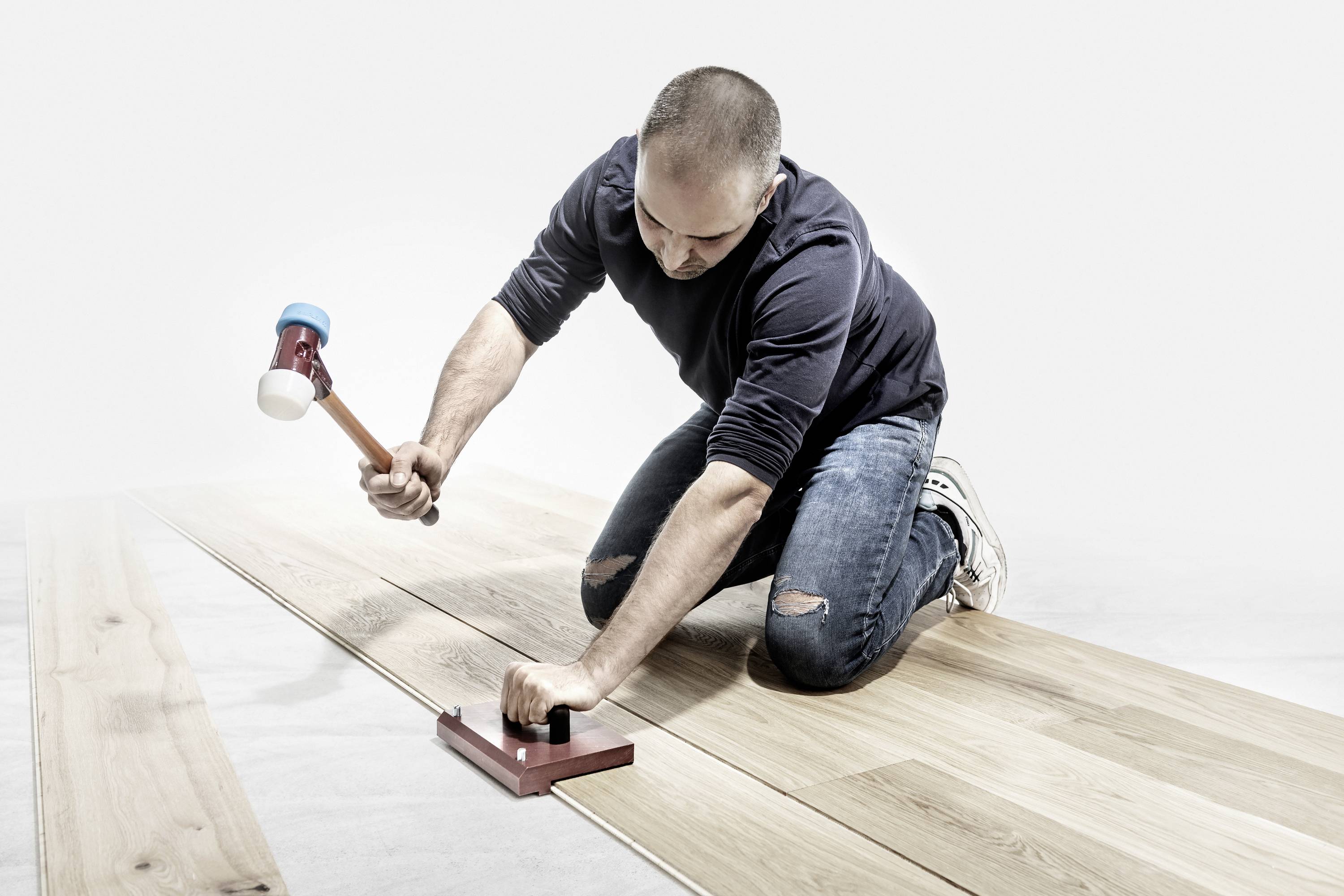 Mężczyzna klęczy i układa podłogę laminowaną. Używa młotka i drewnianego klocka, aby zamocować deski podłogowe.