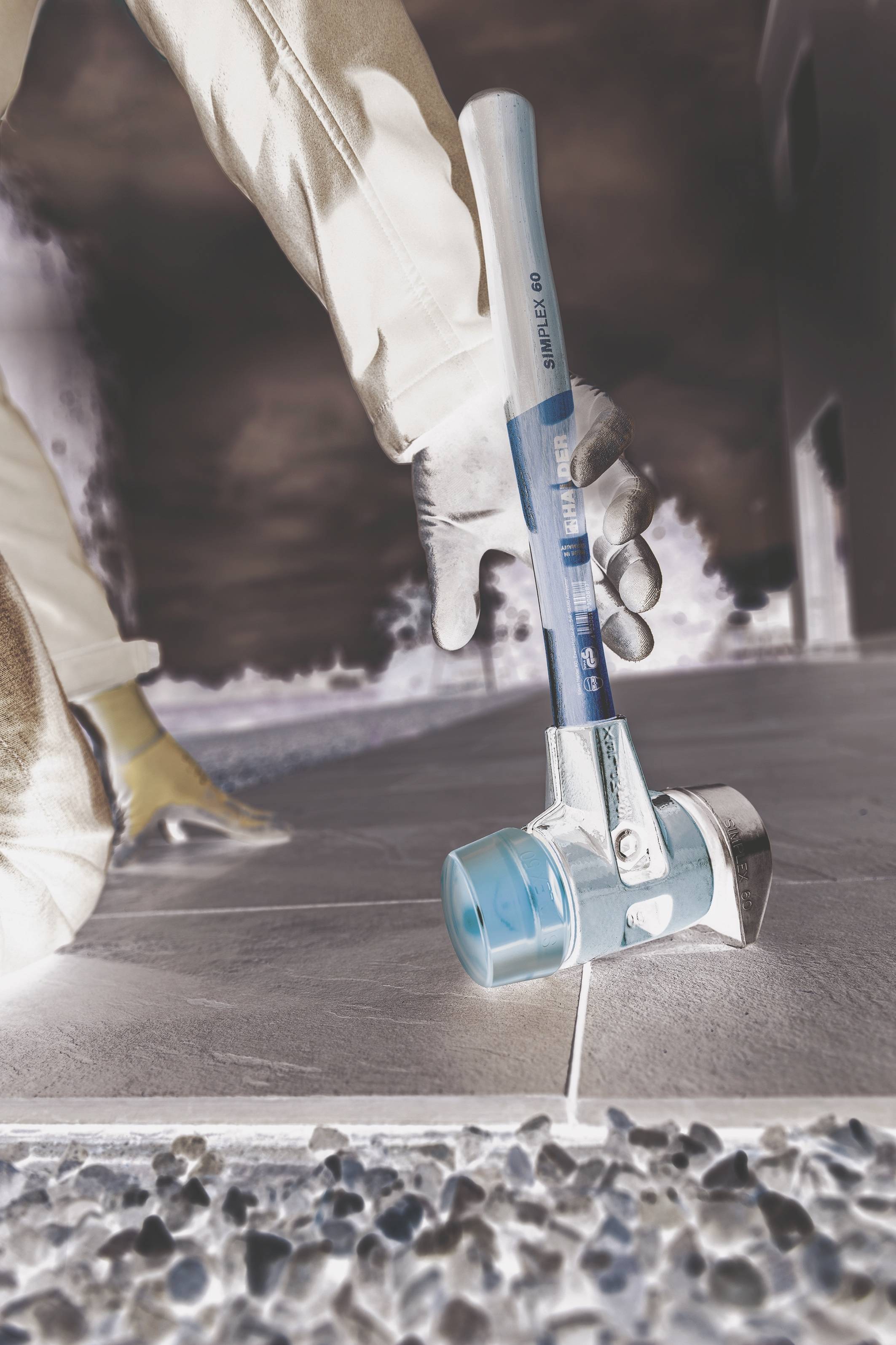 Duży młot z głowicą z tworzywa sztucznego uderzający w płytkę ceramiczną. Osoba w roboczych spodniach i rękawicach klęczy obok tego miejsca na podłodze.