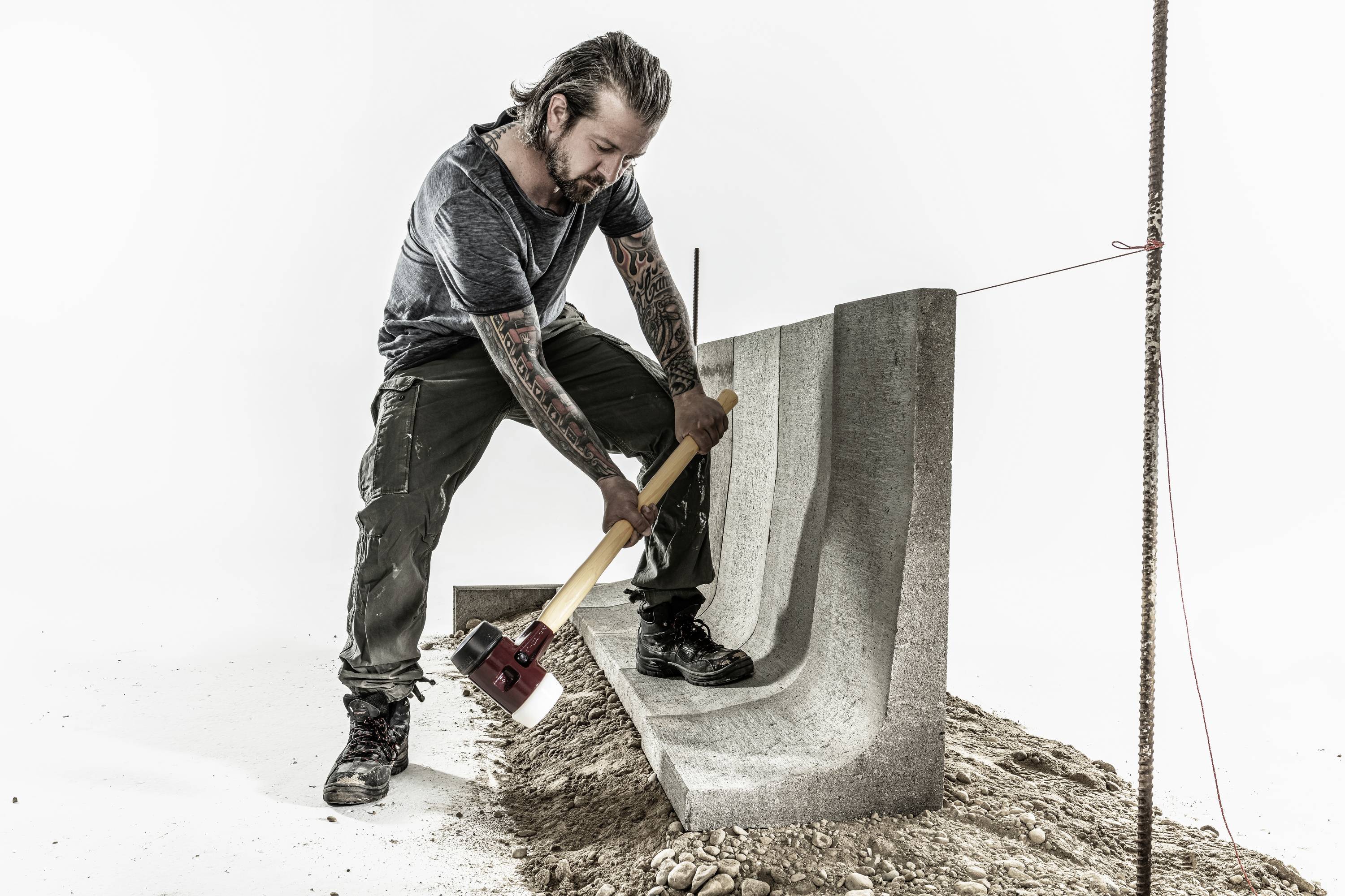 Mężczyzna z tatuażami używa dużego młota do obrabiania bloku betonowego. Ubrany jest w odzież roboczą i stoi na ziemistym gruncie.