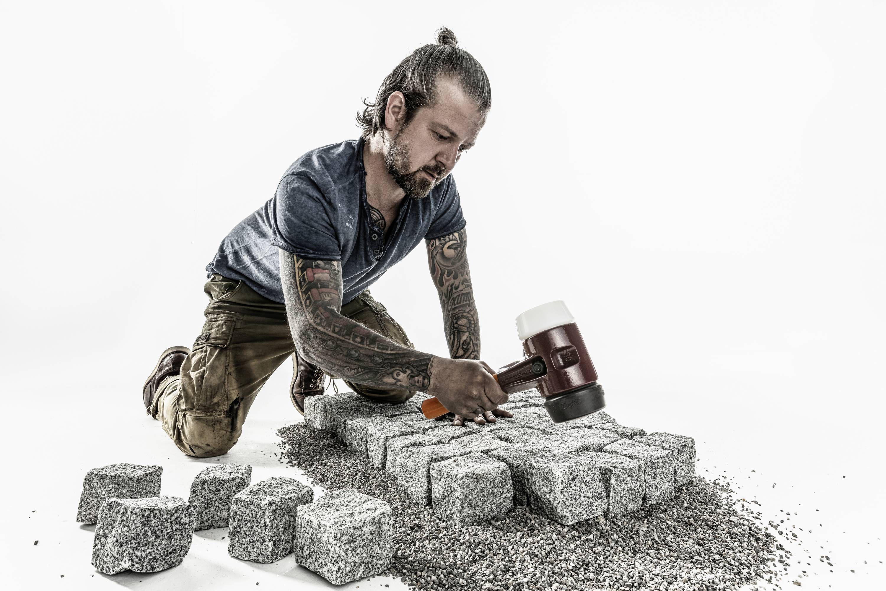 Mężczyzna z tatuażami klęczy na ziemi i układa kostkę brukową. Trzyma młotek gumowy, ma na sobie niebieską koszulę i beżowe spodnie.