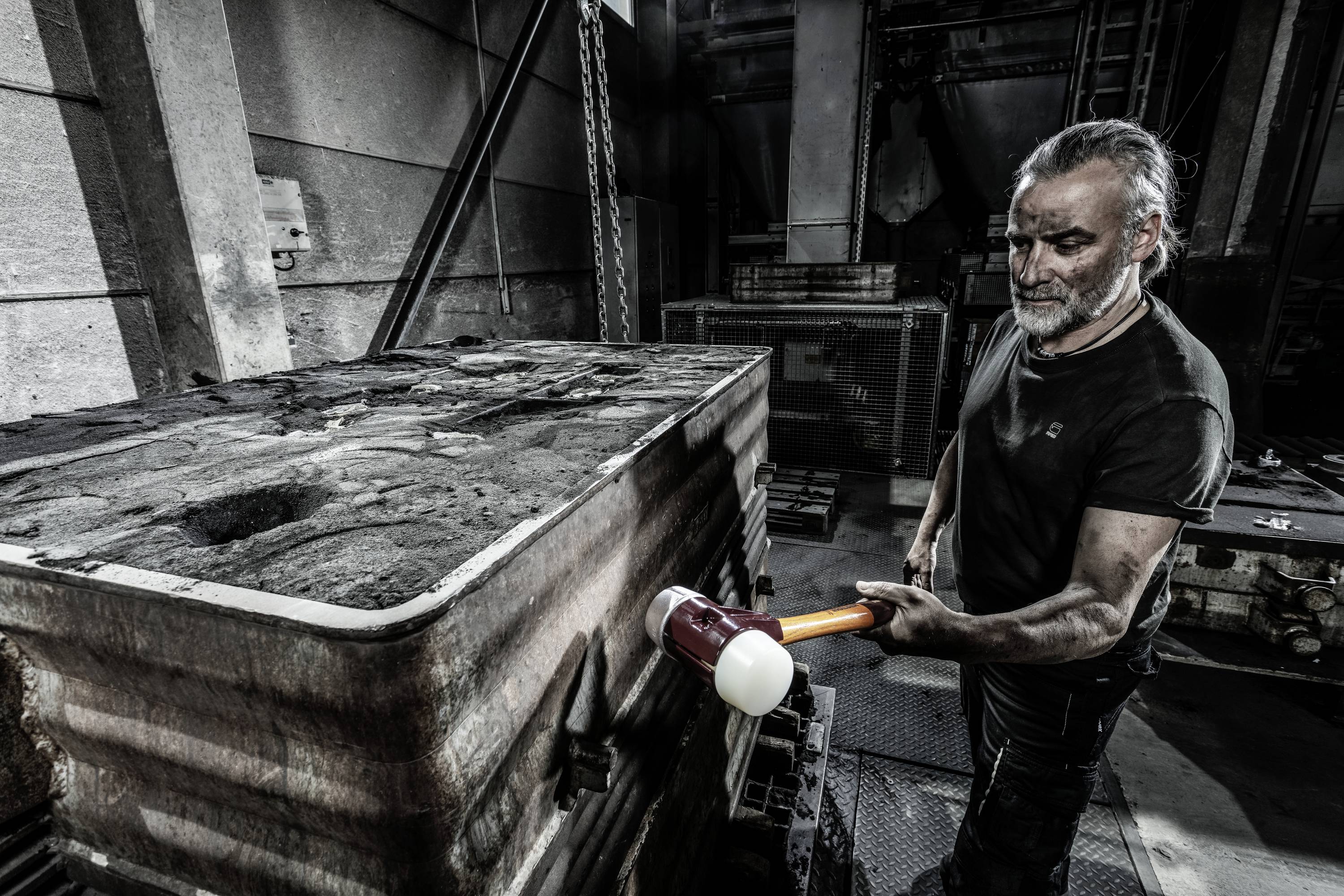 Mężczyzna w ubraniu roboczym stoi w hali przemysłowej i uderza dużym młotem w blok metalowy.