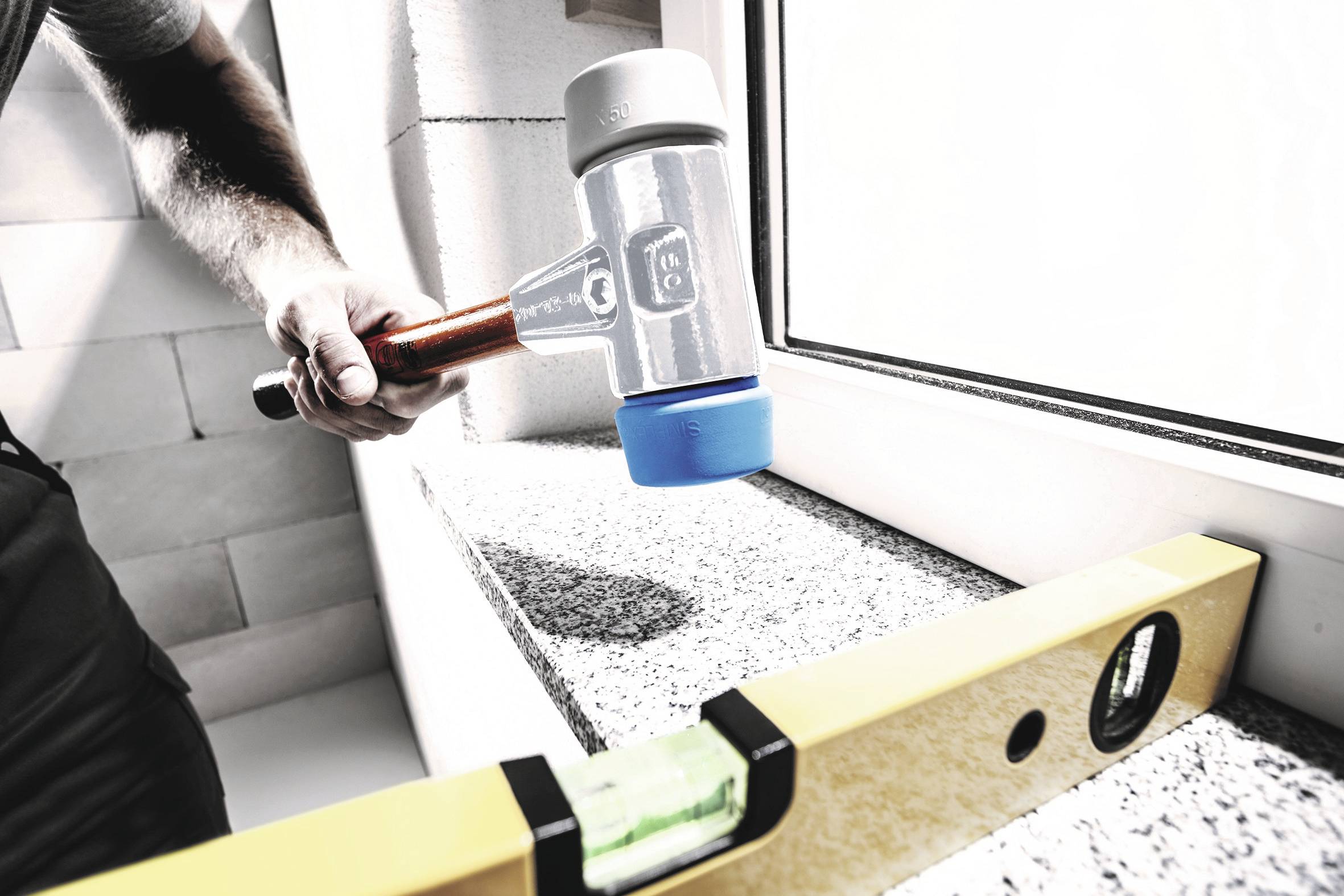 Jedna osoba trzyma młotek gumowy i pracuje przy parapecie. Poziomnica leży na podłożu, sprawdzając równość powierzchni.