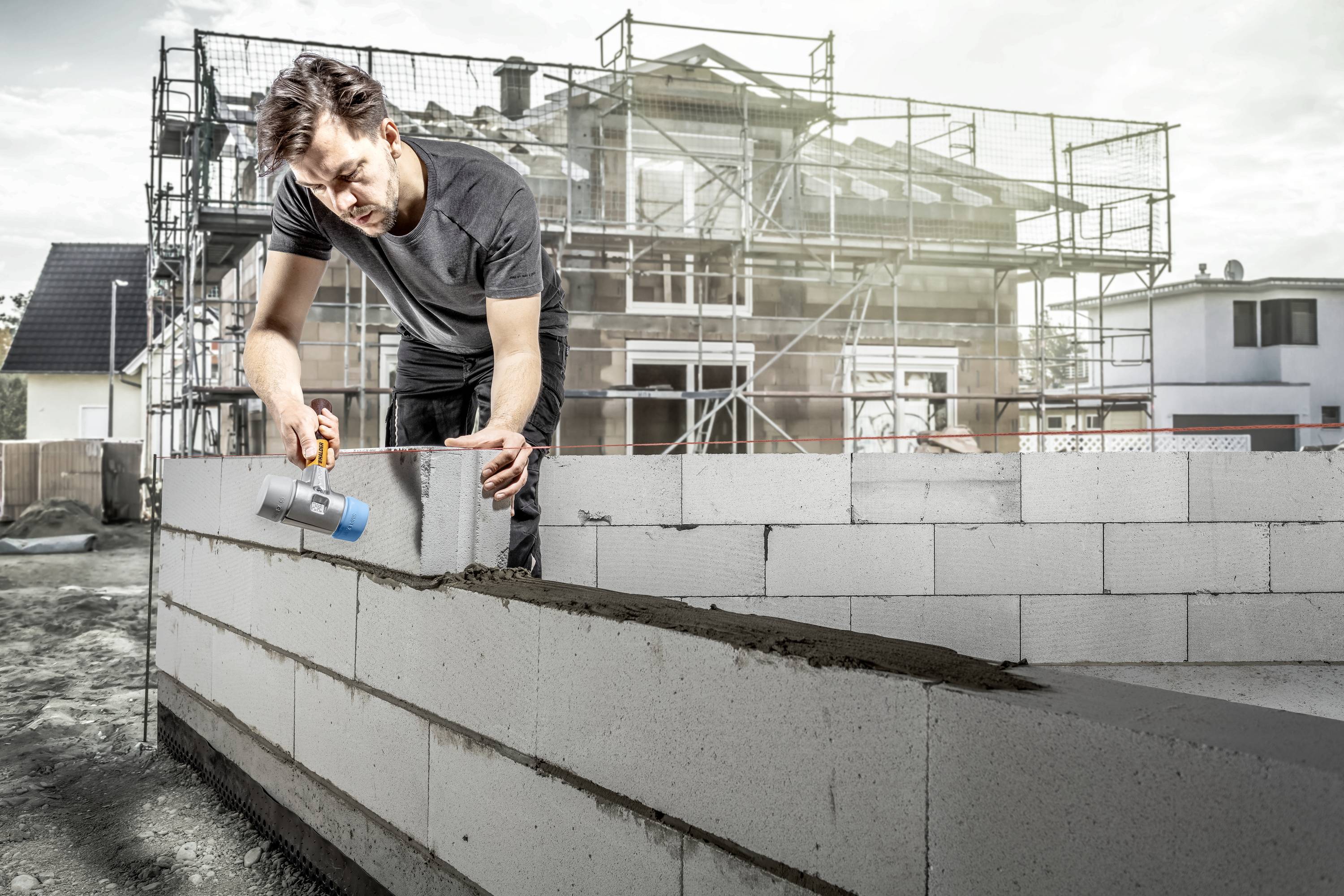 Robotnik budowlany muruje betonowymi blokami na placu budowy przed częściowo ukończonym budynkiem, otoczony rusztowaniami.