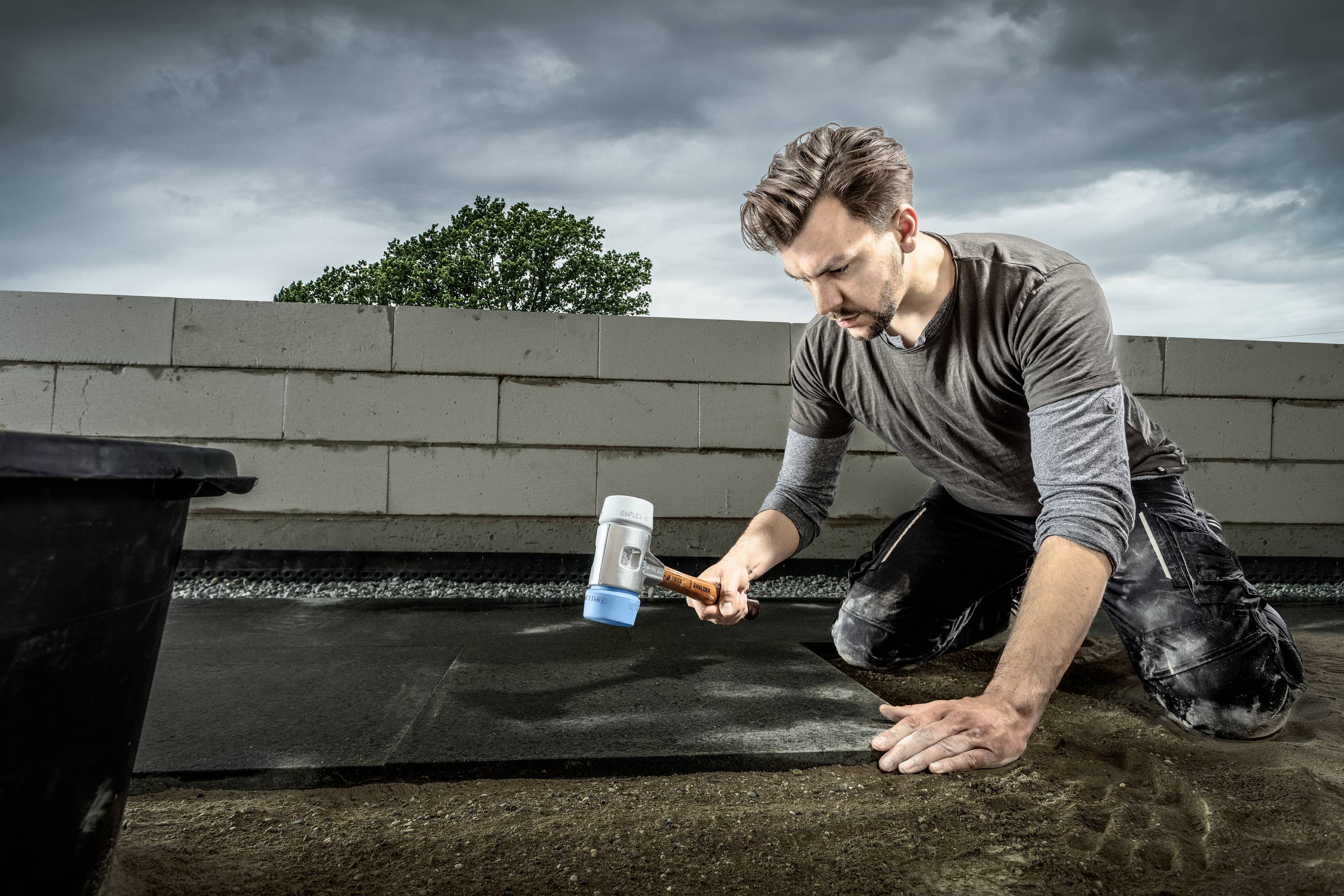 Mężczyzna klęczy na placu budowy i uderza młotkiem w podłogę, w tle widać mur i drzewo.