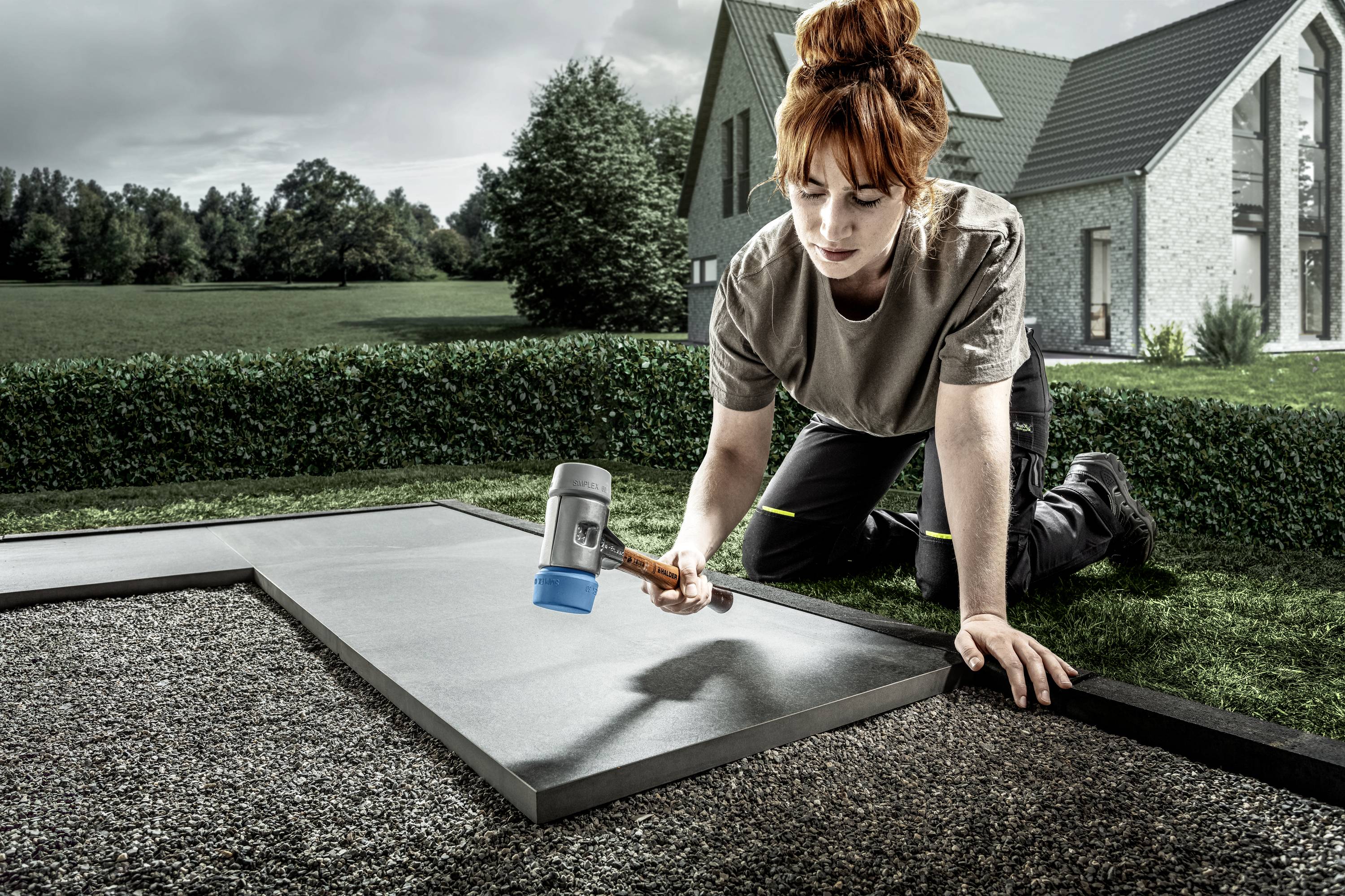 Kobieta z rudymi włosami układa płytę tarasową za pomocą młotka gumowego na podłożu żwirowym w ogrodzie, w tle widać dom.