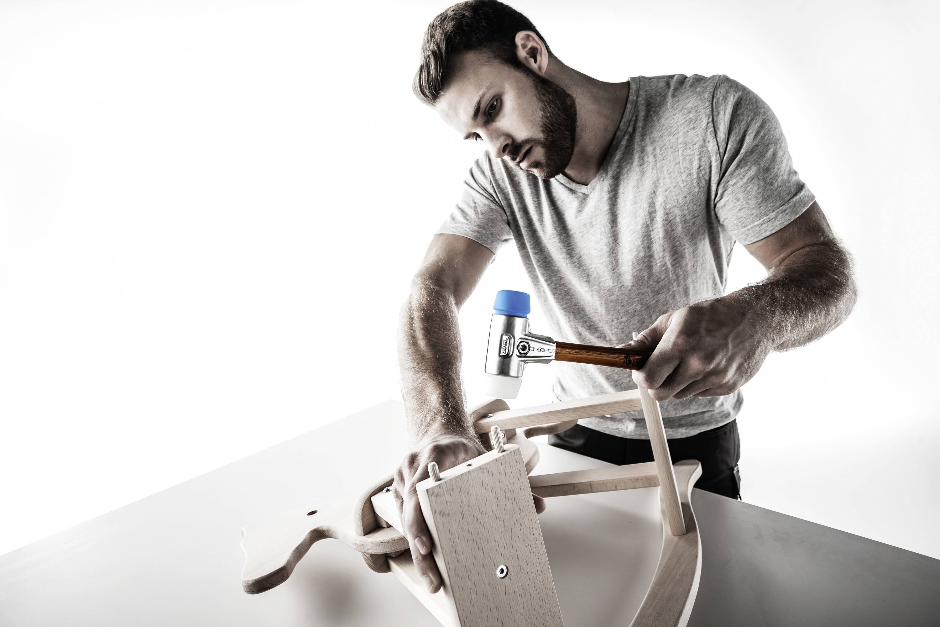 Mężczyzna w szarym t-shircie trzyma młotek i pracuje przy drewnianym krześle na stole.