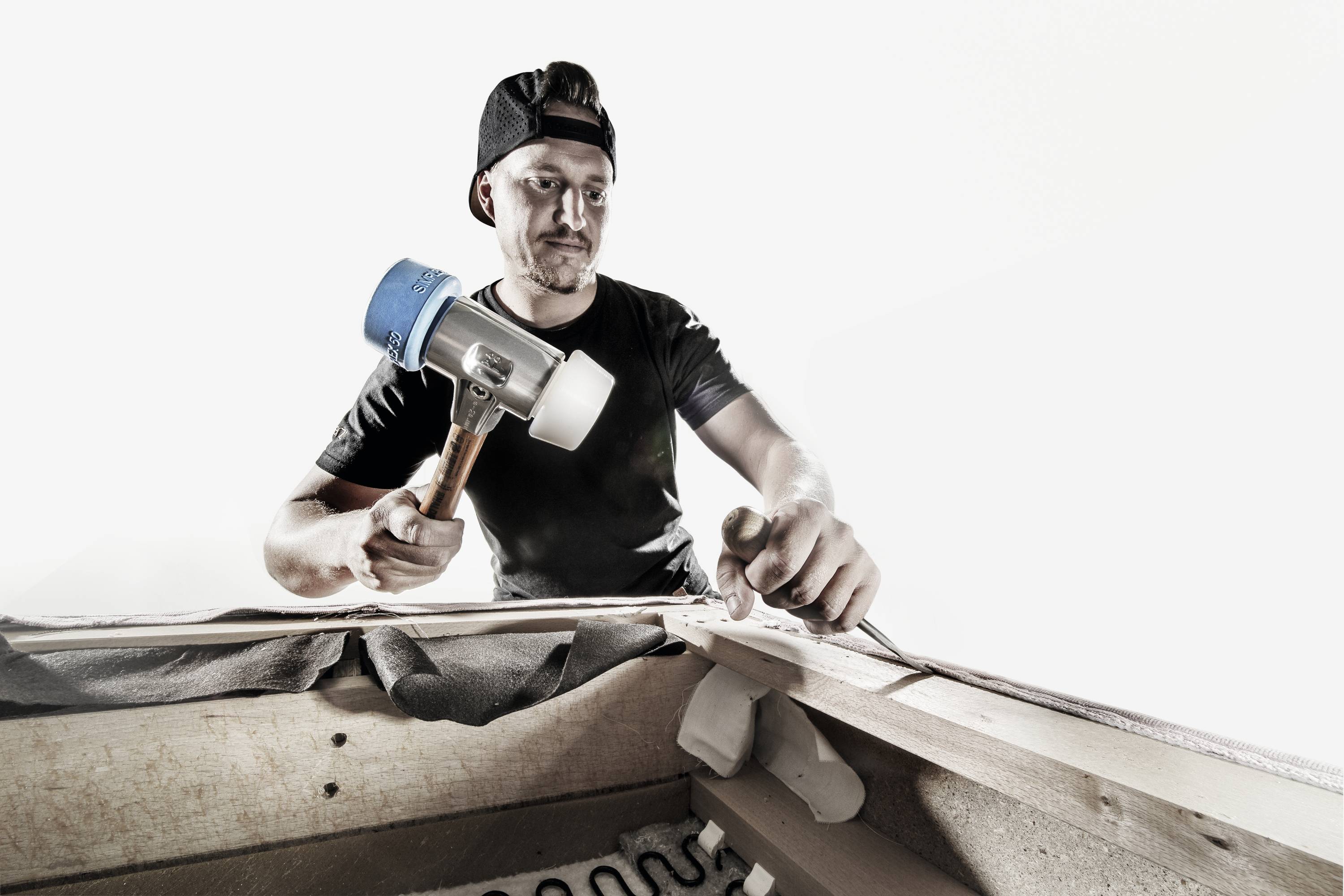 Mężczyzna w czarnym t-shircie i czapce z daszkiem odwróconym do tyłu pracuje narzędziem przy tapicerowanym meblu.