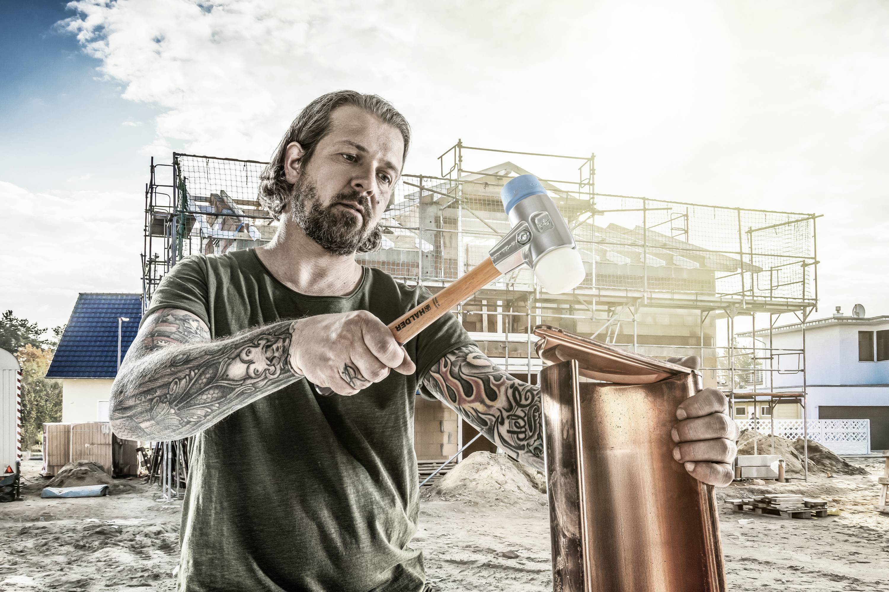 Umięśniony, wytatuowany robotnik budowlany uderzający młotkiem w element miedziany, na tle budowanego domu. Słoneczne światło rozświetla niebo.