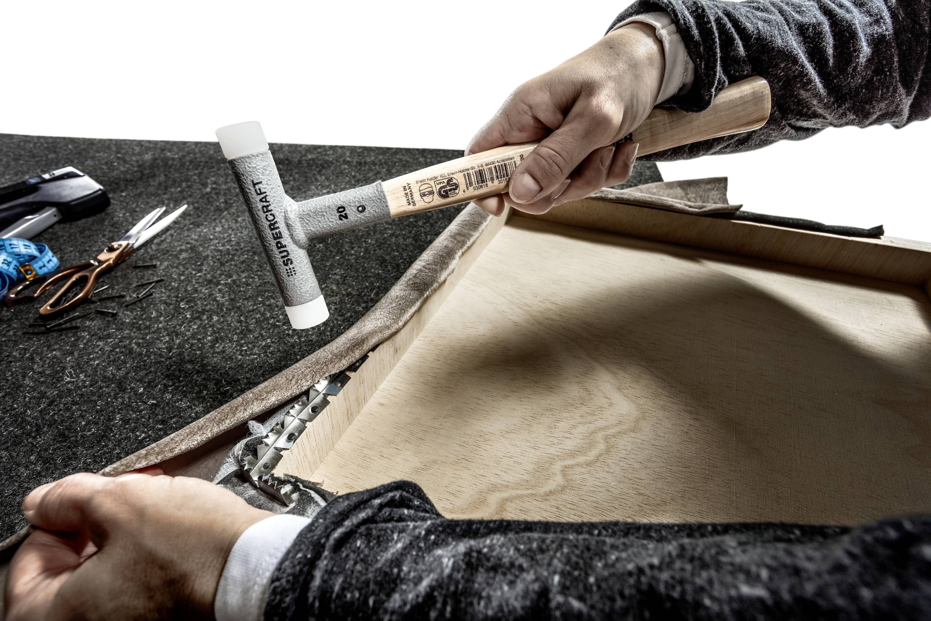 Osoba trzyma młotek gumowy i mocuje tkaninę do drewnianej ramy. Na stole leżą narzędzia i akcesoria.