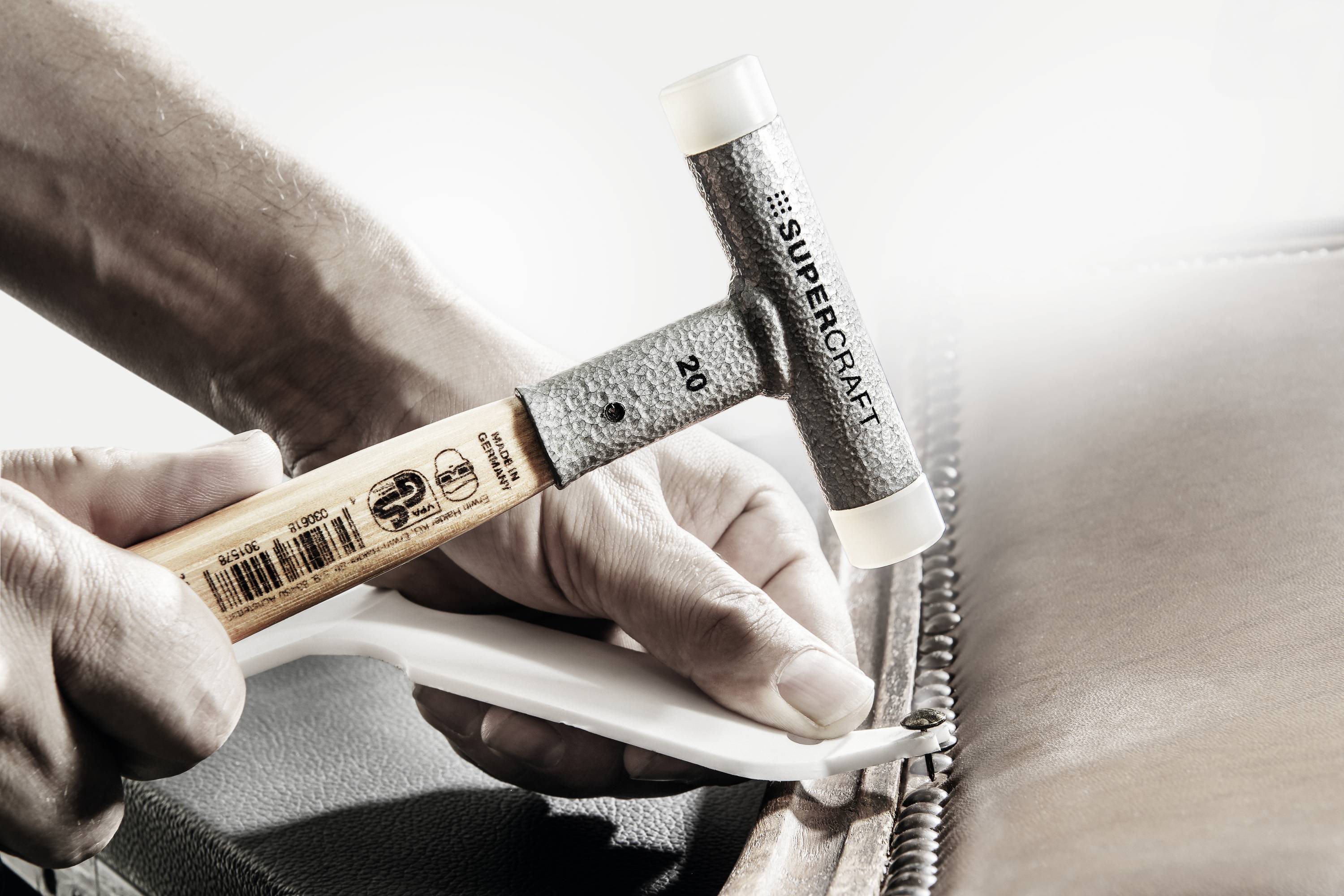 Osoba używa młotka Supercraft do wbijania gwoździ tapicerskich w skórzany mebel. Nacisk kładziony jest na precyzję i rzemieślnicze umiejętności.