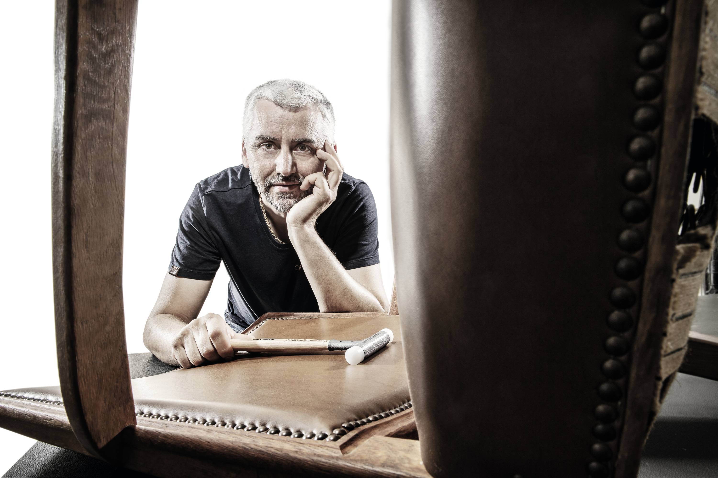 Mężczyzna w czarnym t-shircie zamyślonym wzrokiem przyglądą się skórzanej powierzchni przewróconego krzesła, obok leży miarka krawiecka.