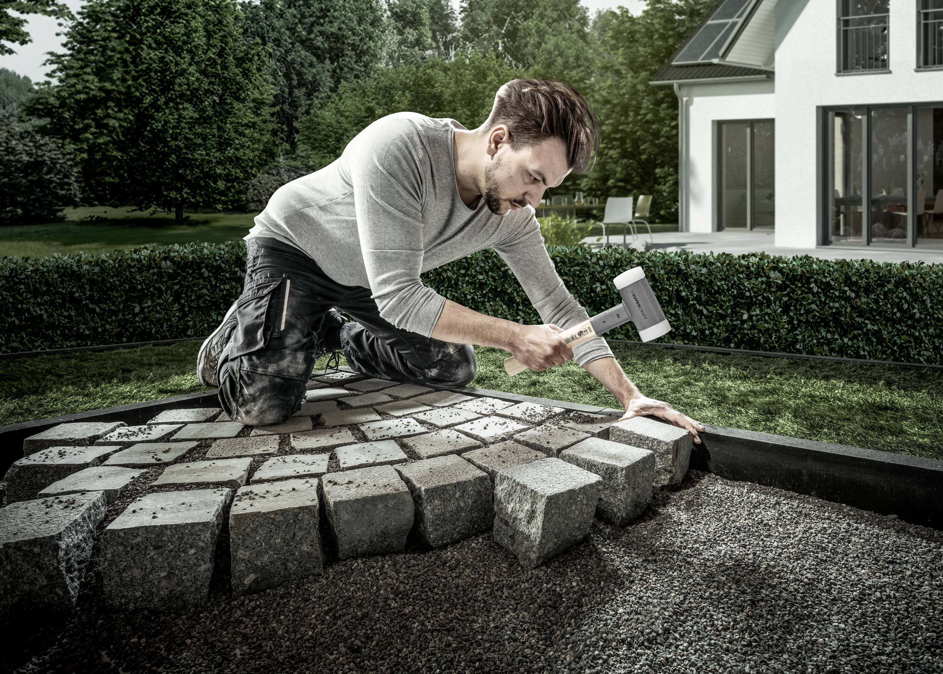 Mężczyzna klęczy na ścieżce ogrodowej i wbija kostki brukowe młotkiem. W tle widać nowoczesny dom.
