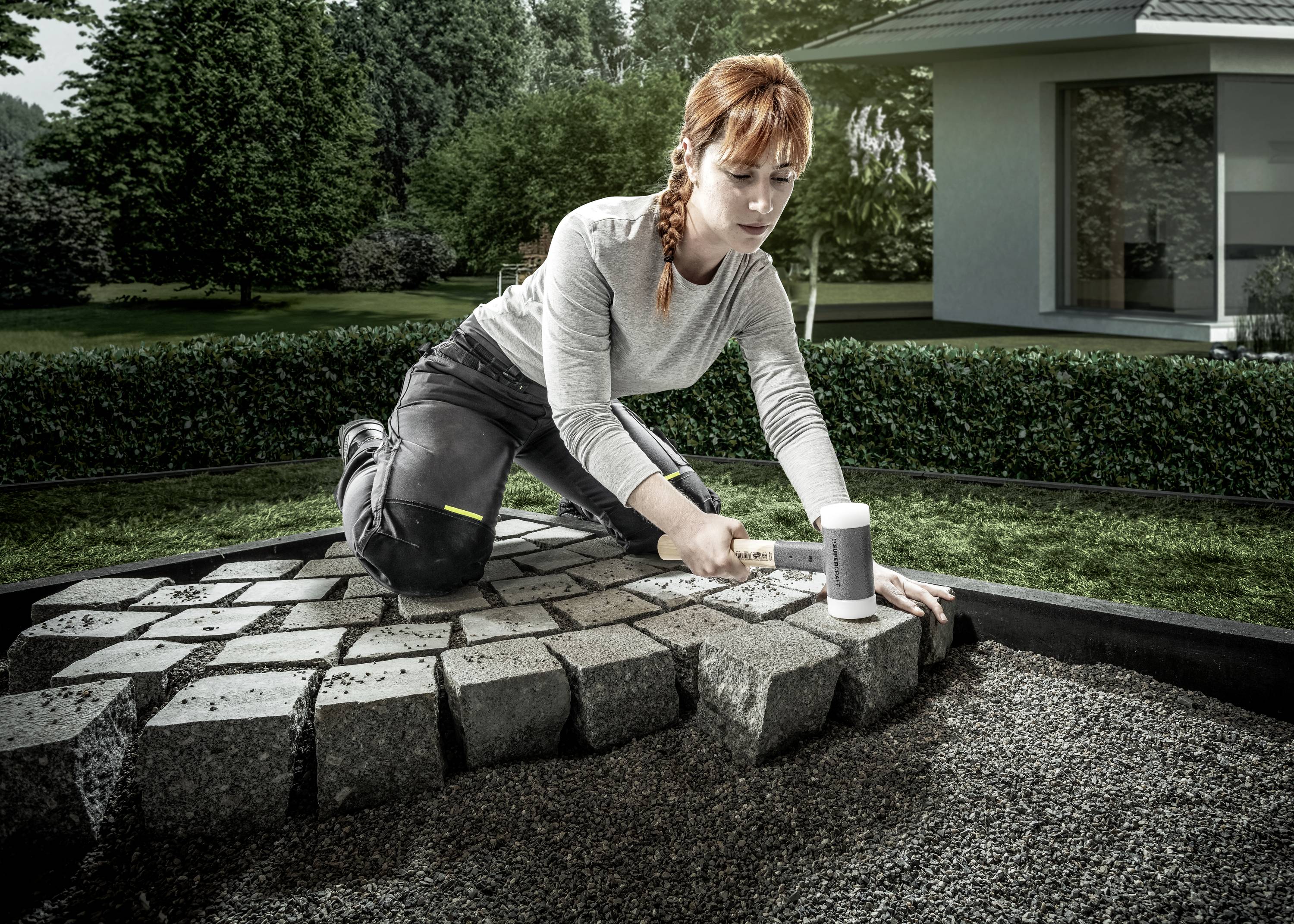 Osoba układa kamienne płyty na tarasie lub chodniku w ogrodzie. W tle widać nowoczesny dom.